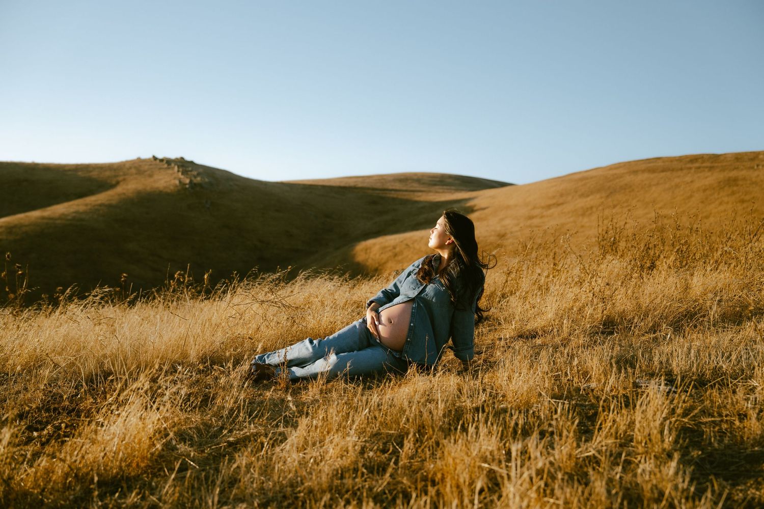 A woman in jeans and jacket sits relaxed on a golden hillside meadow under a clear blue sky.