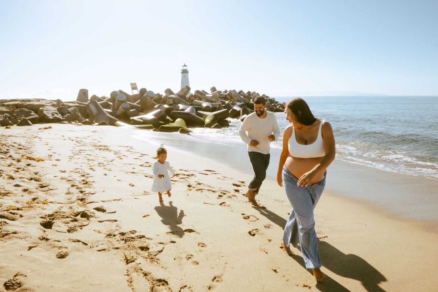 Family walking on sunny beach near rocky jetty with lighthouse, parents and child enjoying seaside stroll.