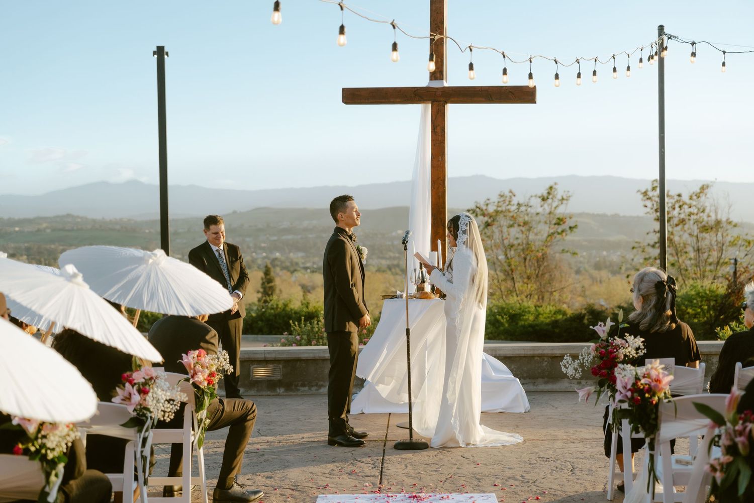 Outdoor wedding ceremony at a hilltop venue with a wooden cross, string lights, mountain views, and floral arrangements.