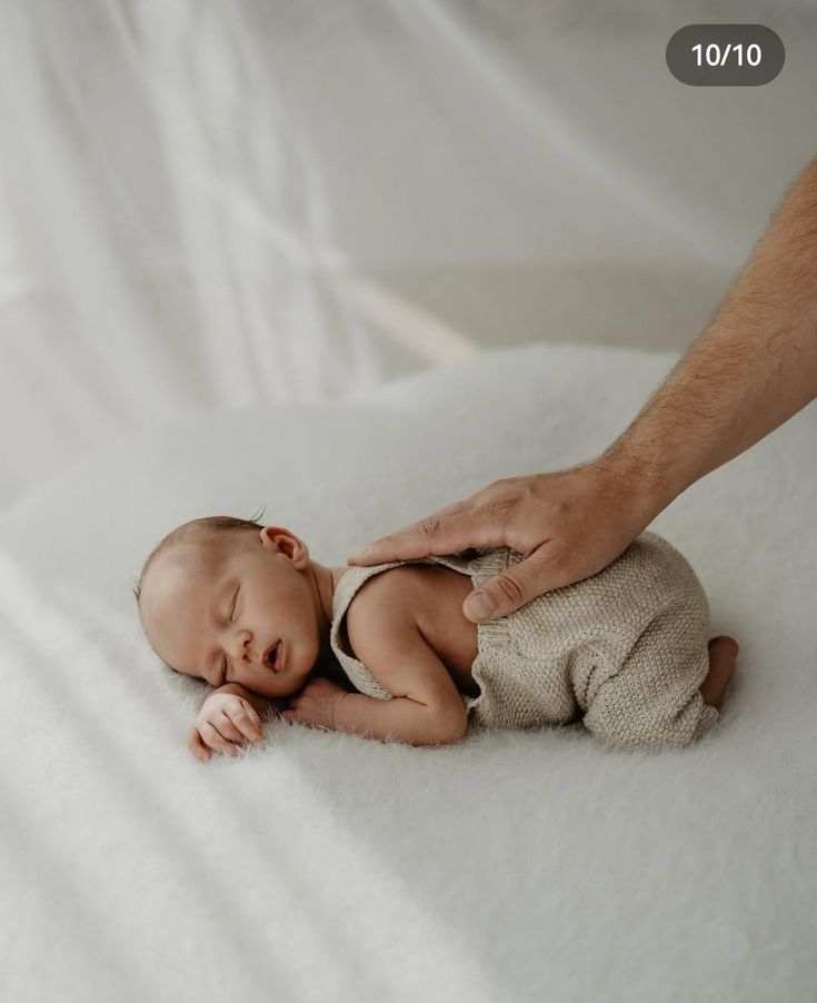 Gentle hand reaches out to comfort sleeping newborn baby wrapped in soft gray blanket on white backdrop.