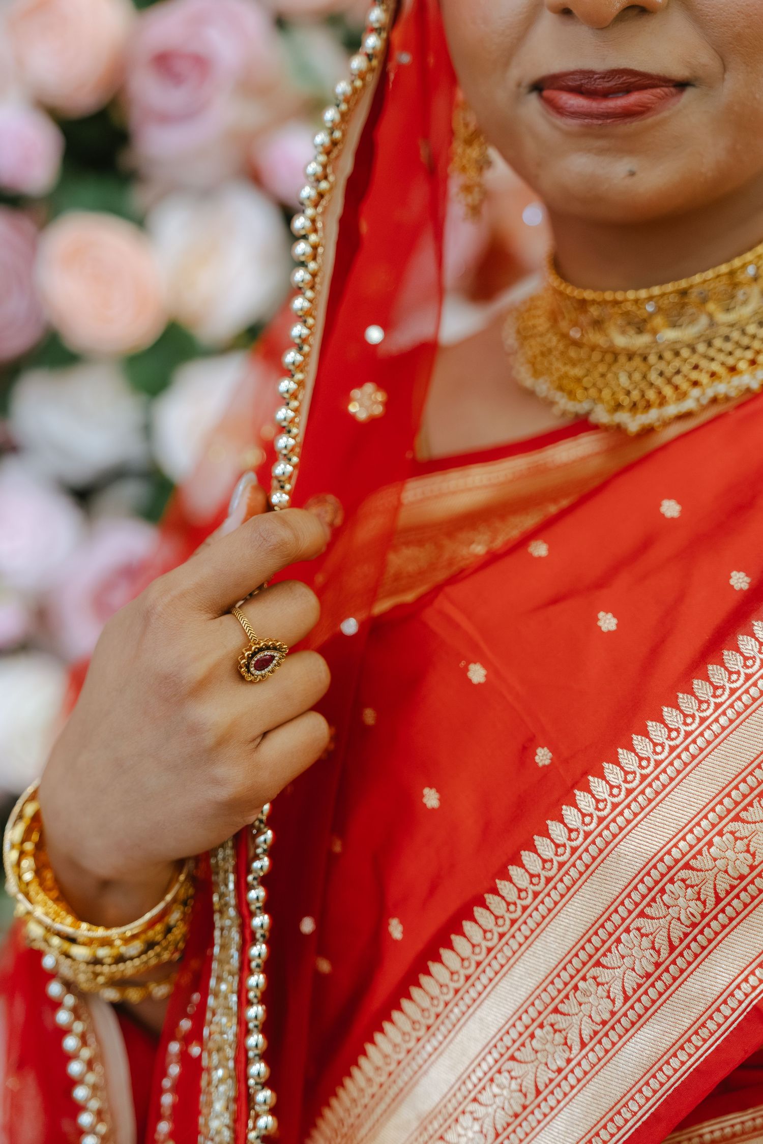 Indian bride in red saree with gold jewelry, bangles, and ring holding her dupatta near floral backdrop.