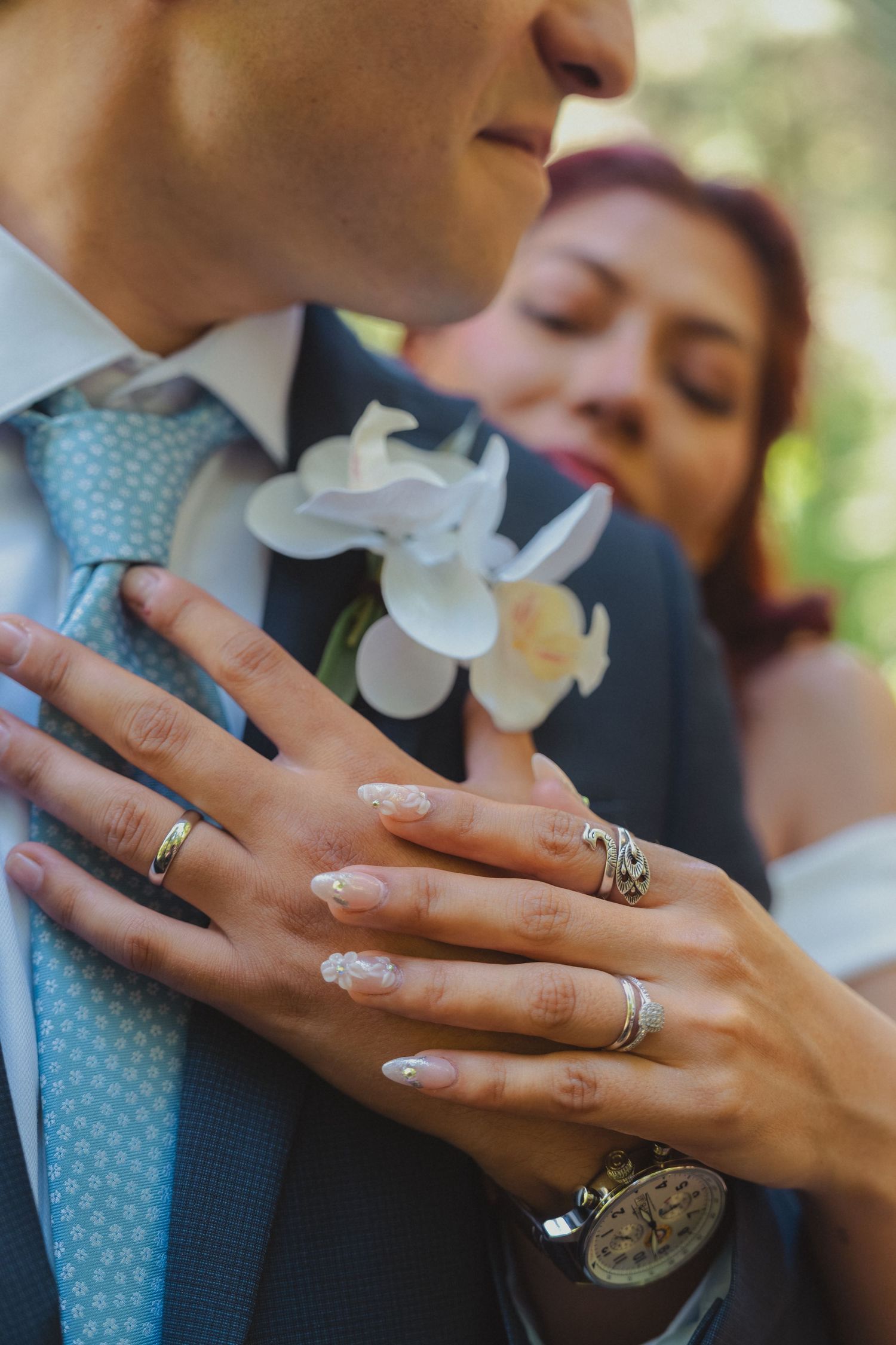 Newlyweds displaying wedding rings on grooms chest near white orchid boutonniere and blue tie.