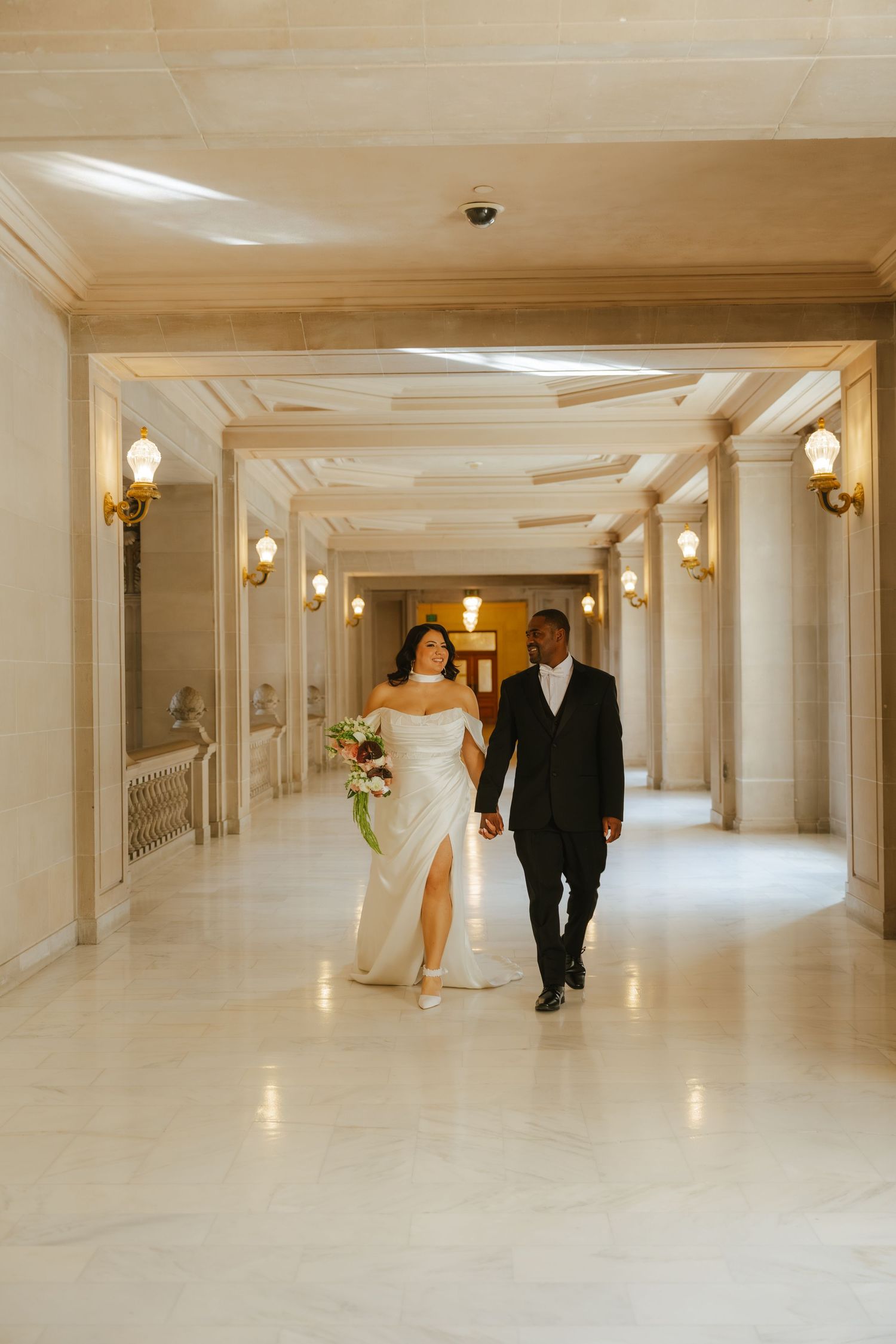 Bride and groom walk hand-in-hand through elegant marble hallway, bride in white gown holding bouquet.