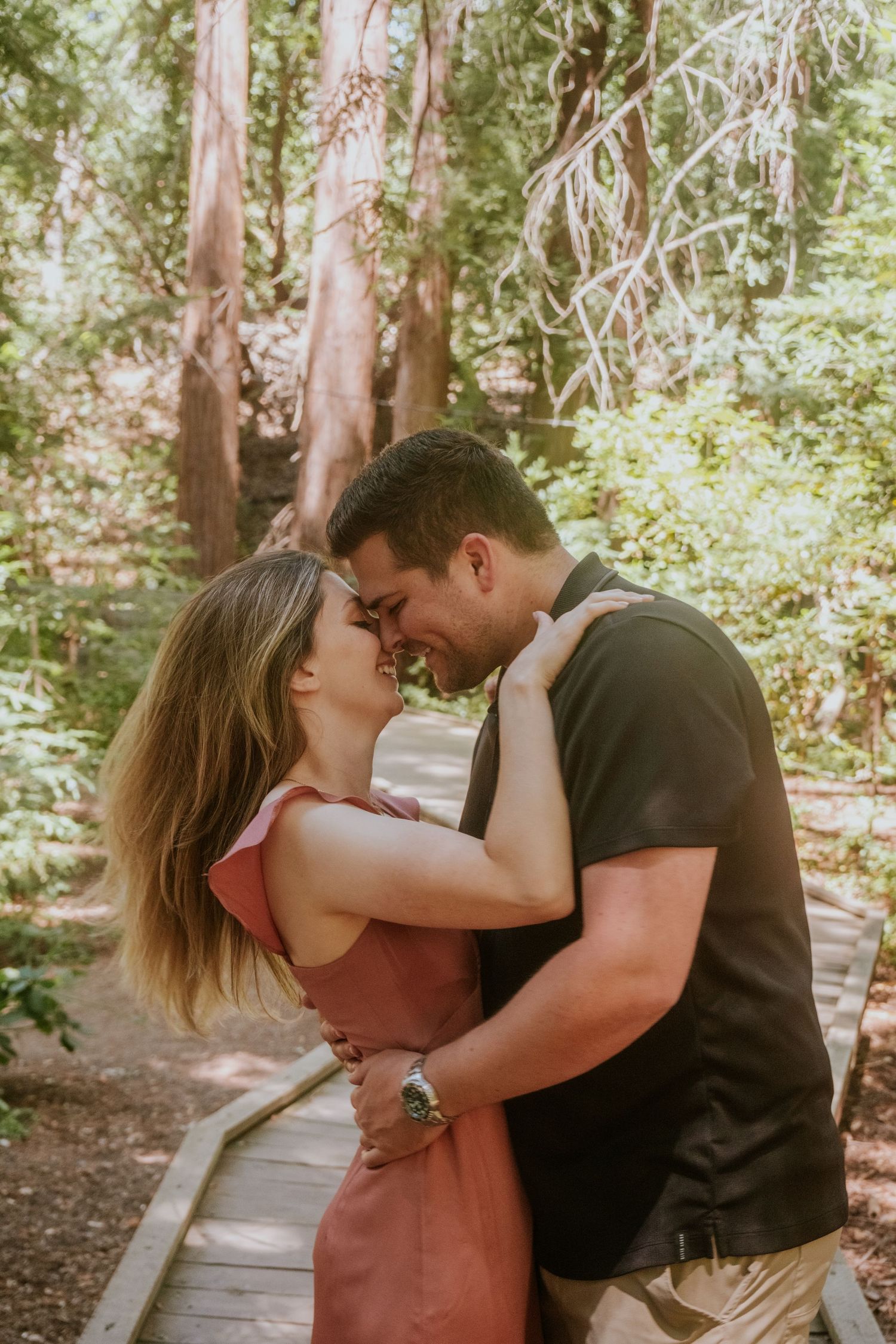 Intimate couple moment captured on forest boardwalk with filtered sunlight through trees.