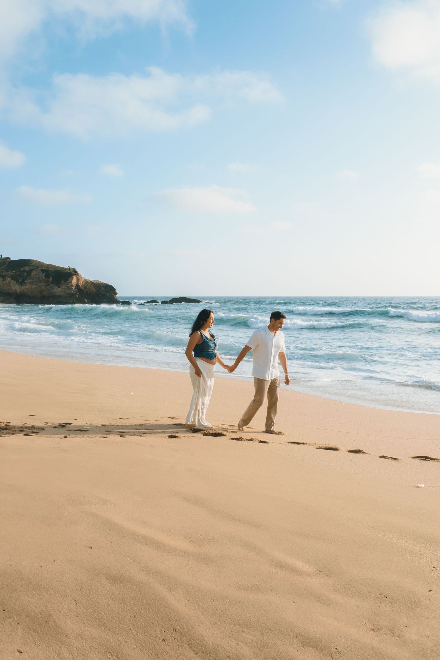 A couple walks together on a sandy beach with ocean waves and cliffs in the background.