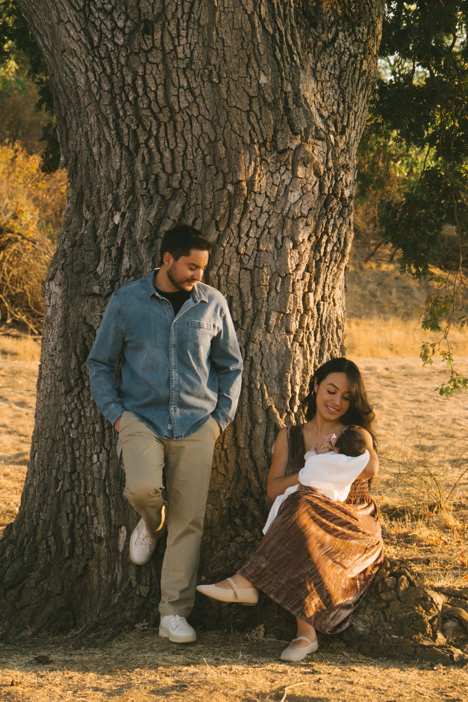 Family sitting together against large tree trunks in golden afternoon light.
