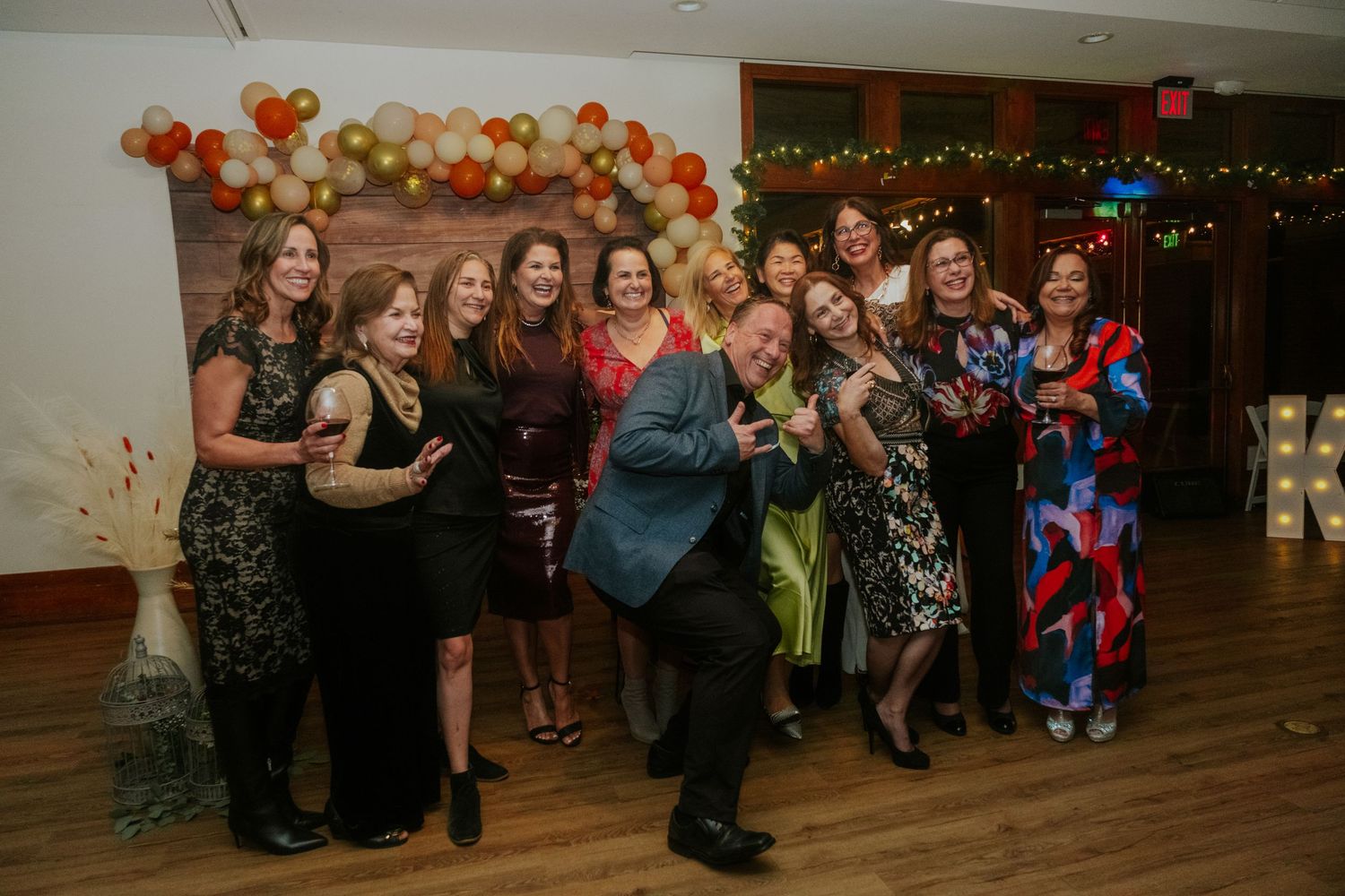 Group of women and one man posing and laughing at a festive party with balloon arch backdrop.