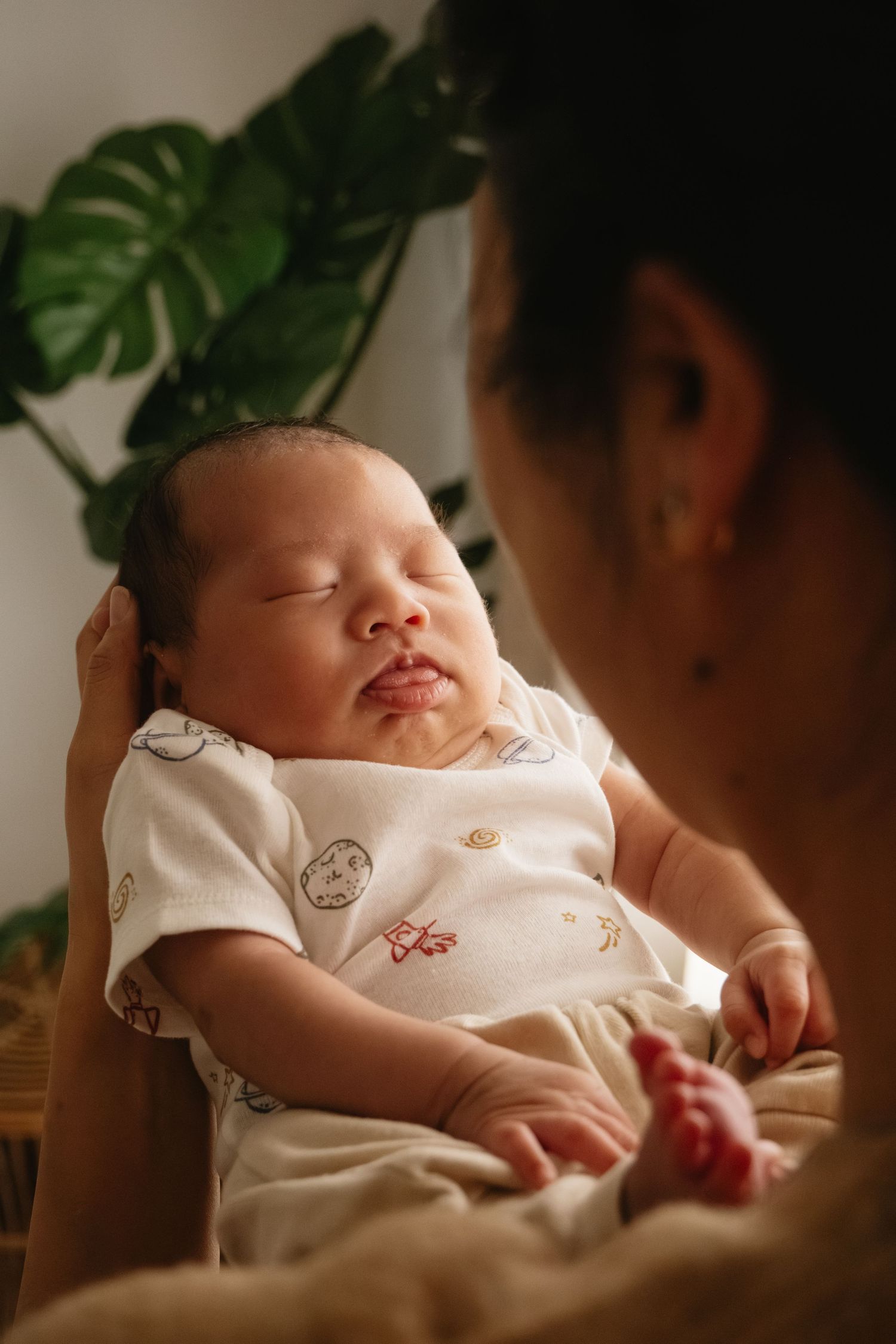 A newborn baby sleeping peacefully in a parent's arms, wearing a white onesie with small prints.