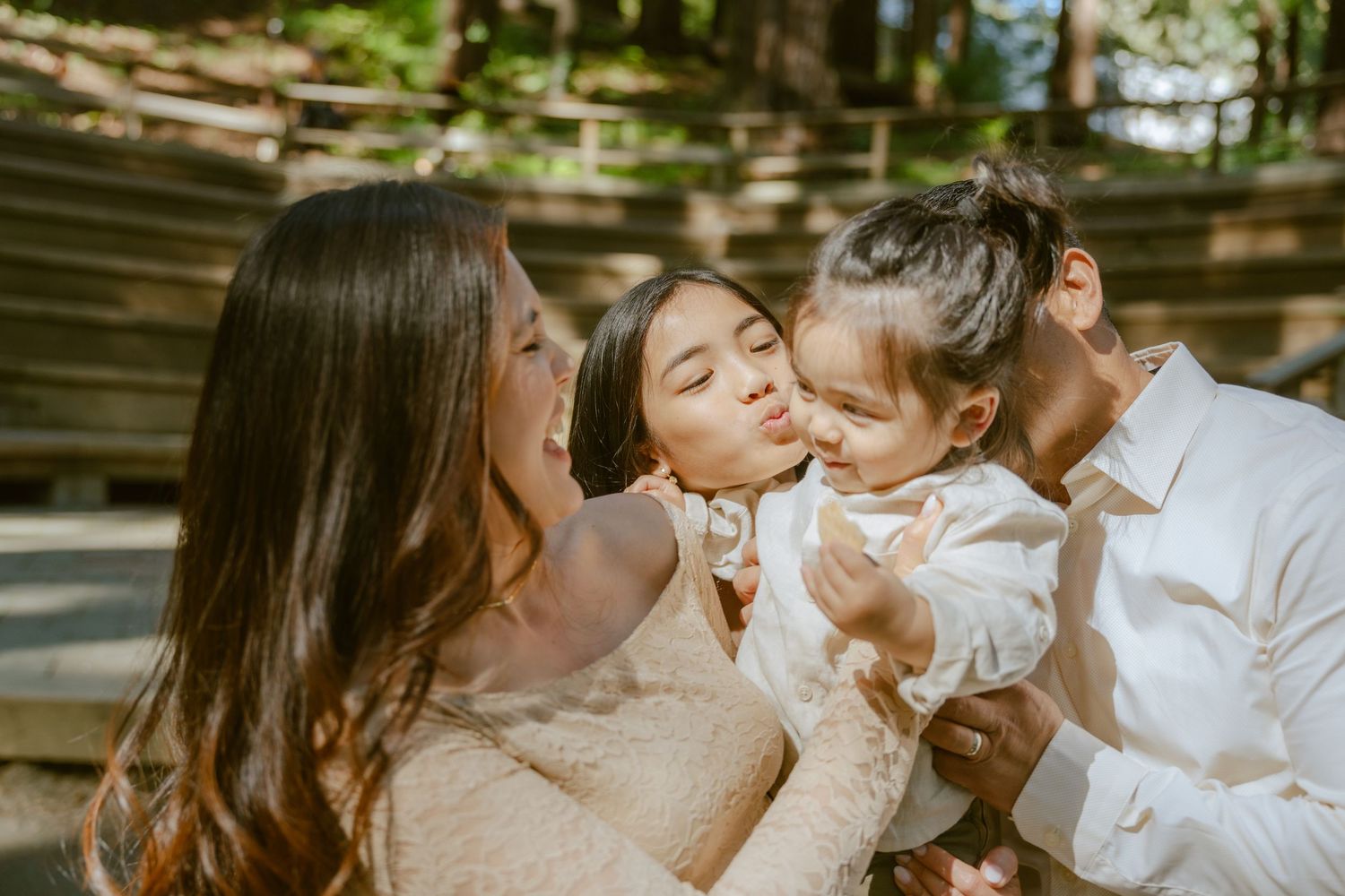 Family photo with parents and two young children sharing kisses outdoors in a sunlit wooded setting.