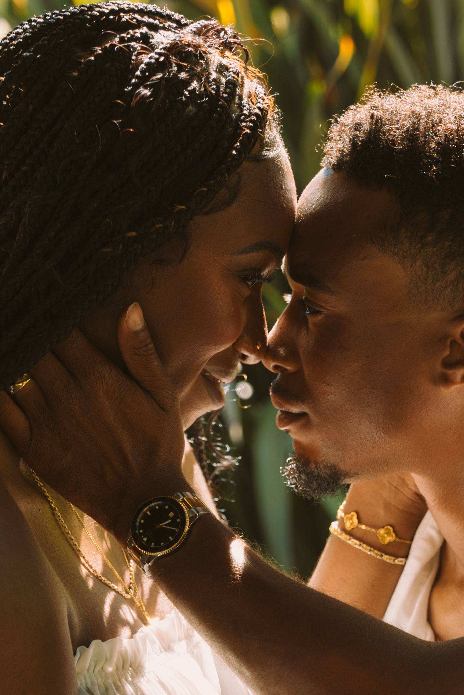 Two people sharing an intimate moment, foreheads touching, wearing gold jewelry and a watch in warm sunlight.