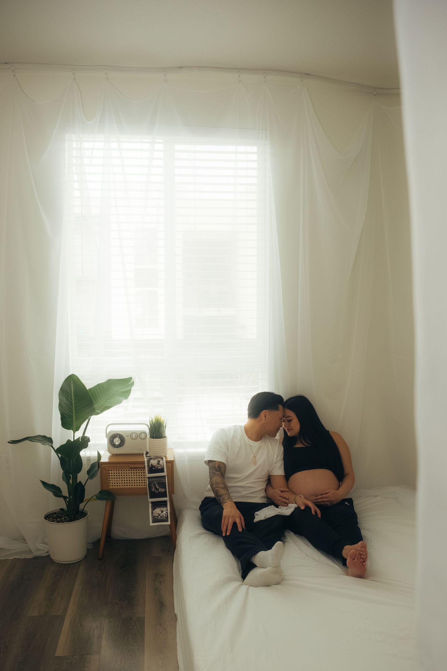 Minimalist white bedroom interior with houseplants and natural light streaming through sheer curtains.