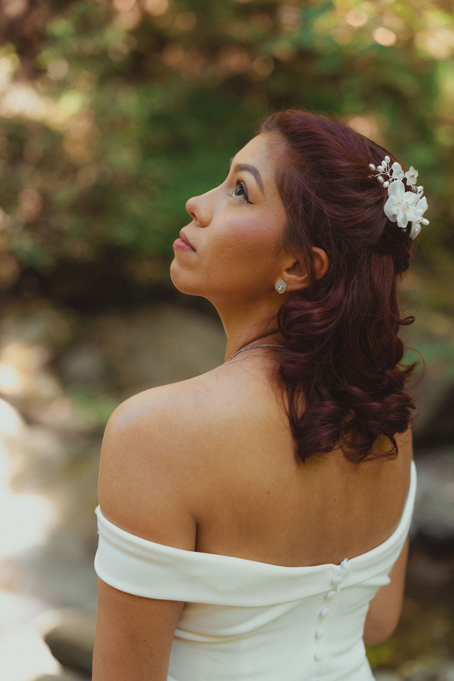 Bride in off-shoulder white gown with white floral hair accessory and red hair gazing upward outdoors.