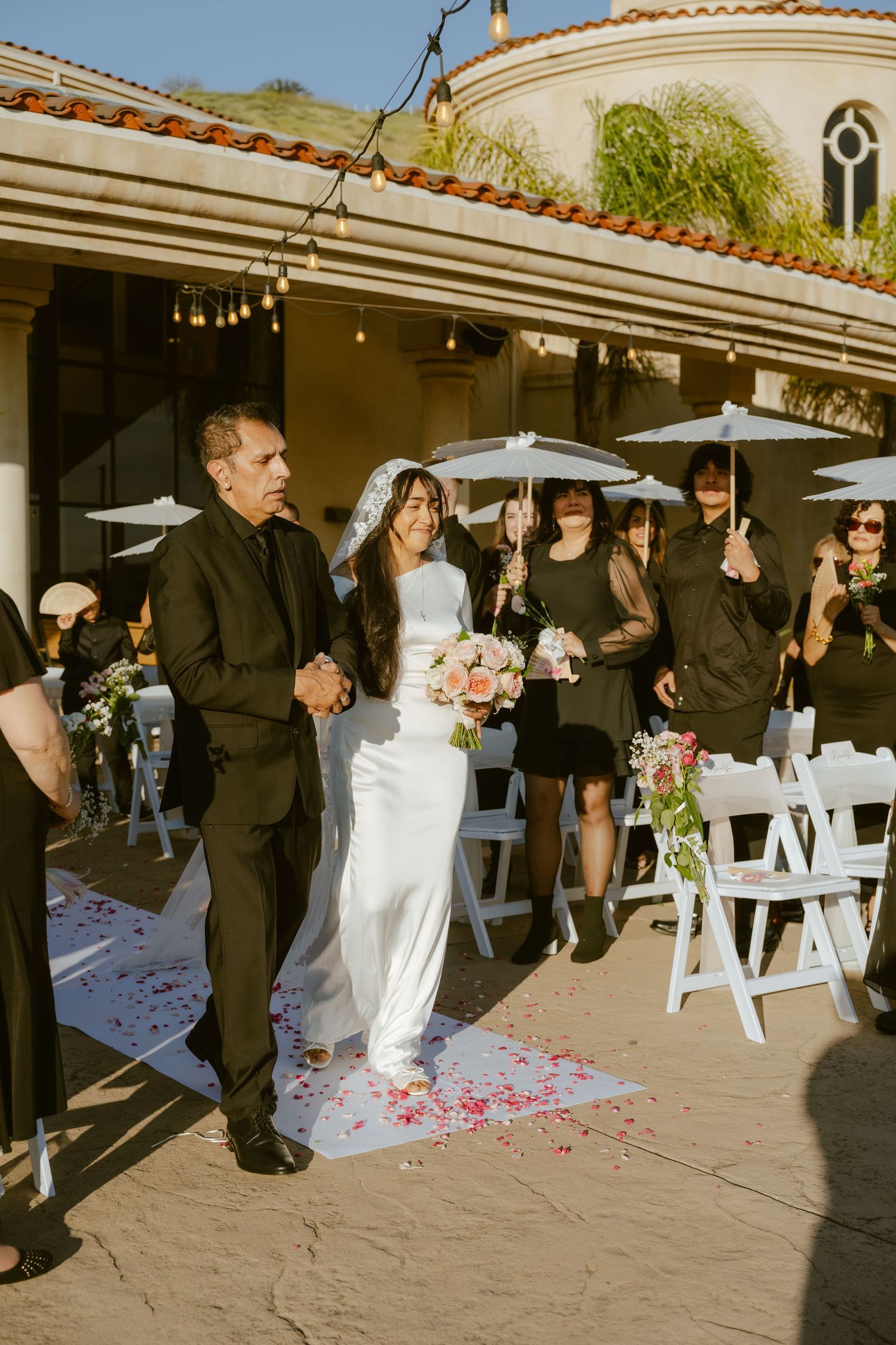 Bride in white gown walks down outdoor aisle with guests holding umbrellas at a sunny wedding ceremony.