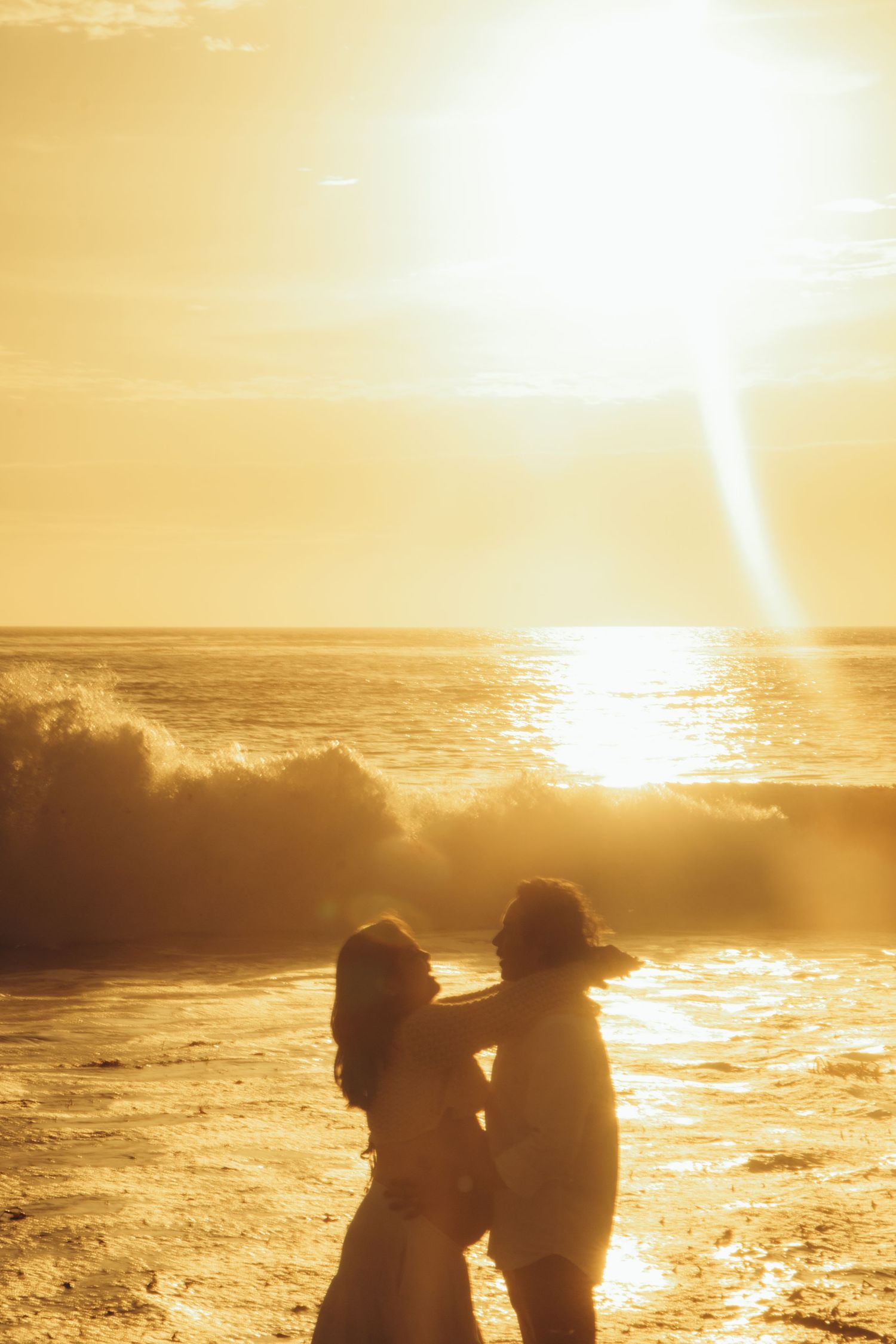 Silhouettes of people embracing on beach during golden sunset with waves crashing.