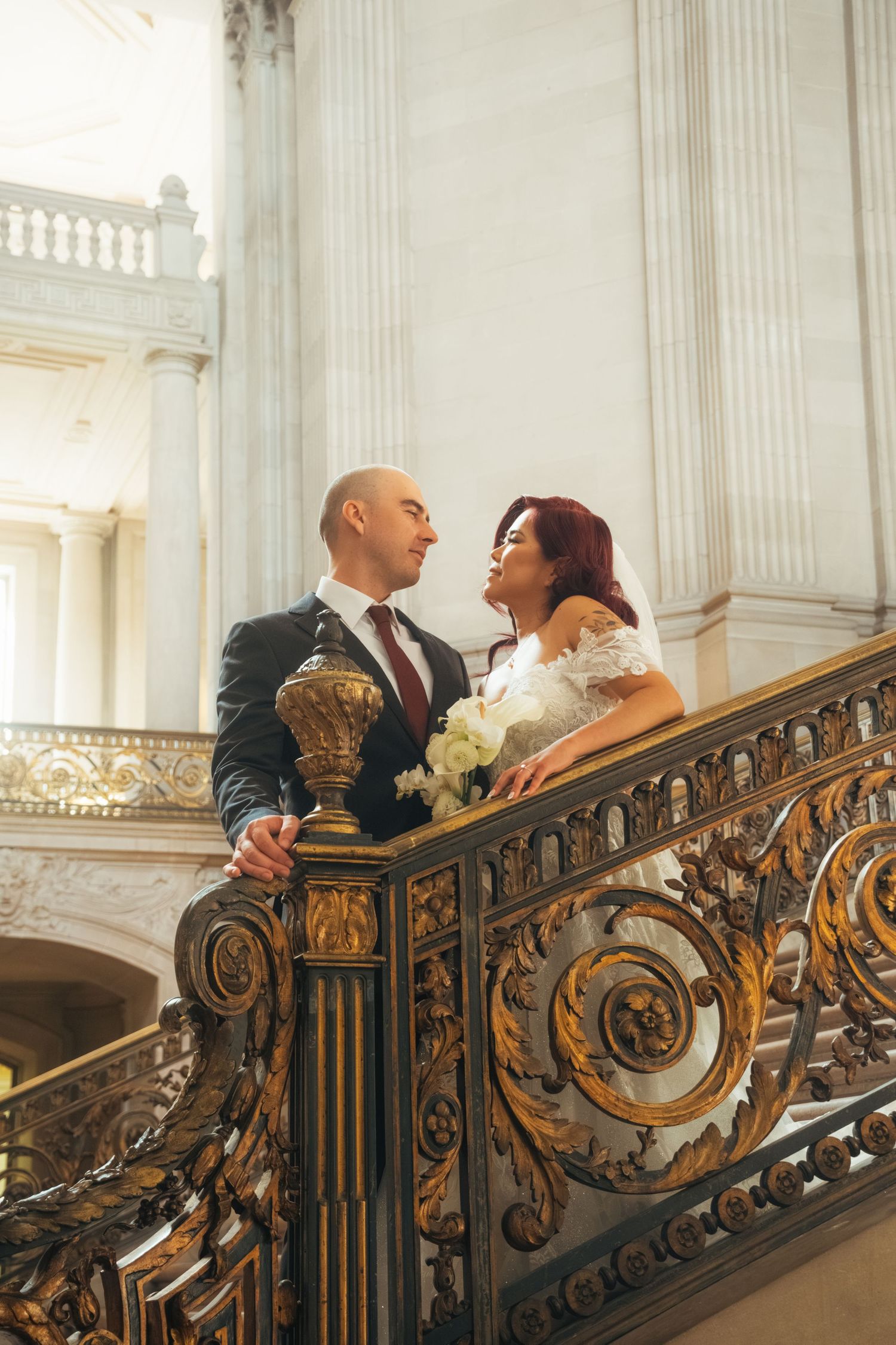 Wedding couple embraces by an ornate gold railing in a historic building with classical architecture.