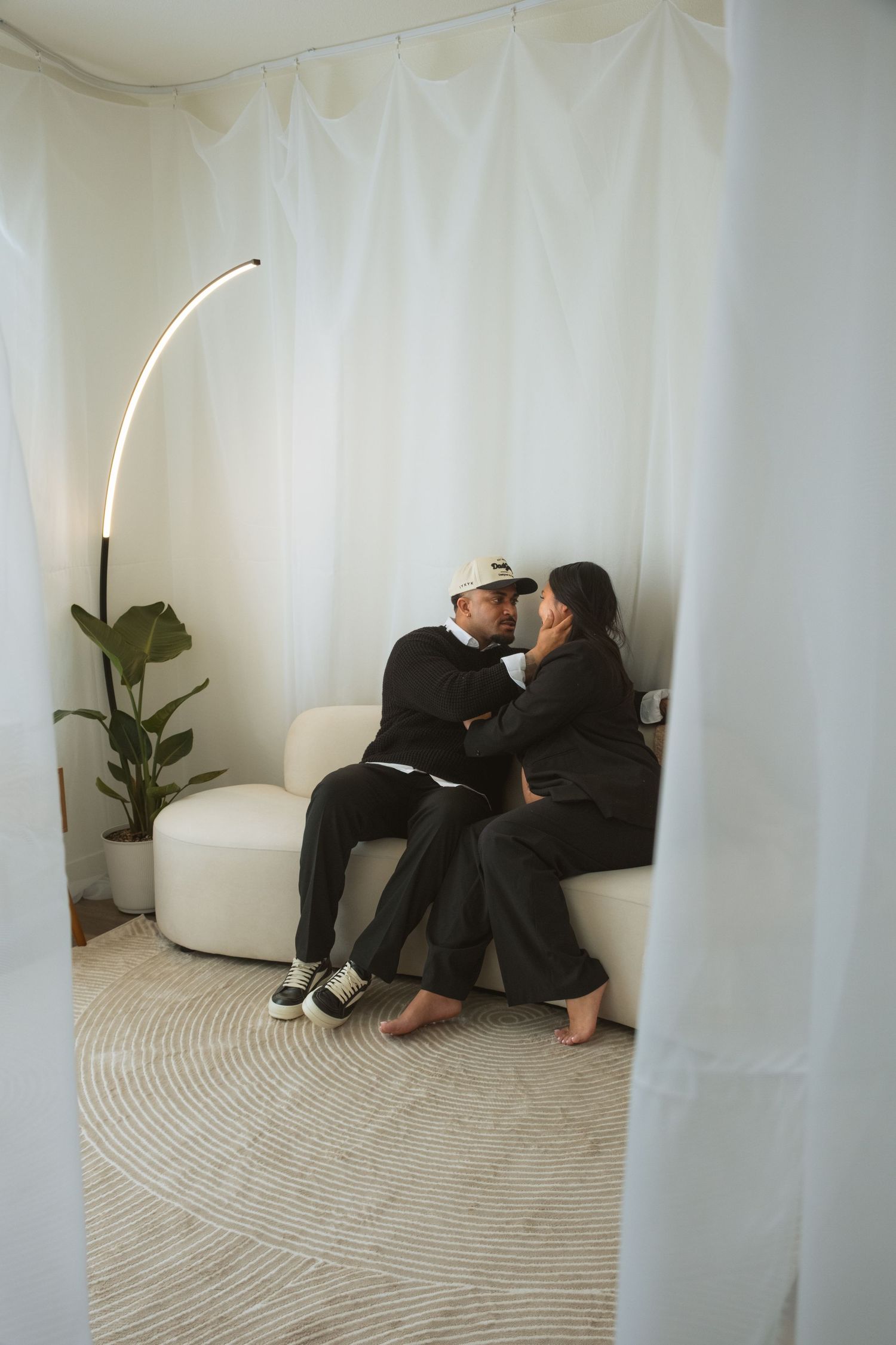 Two people in dark clothing sit intimately on white furniture in a minimalist white room with curved walls.