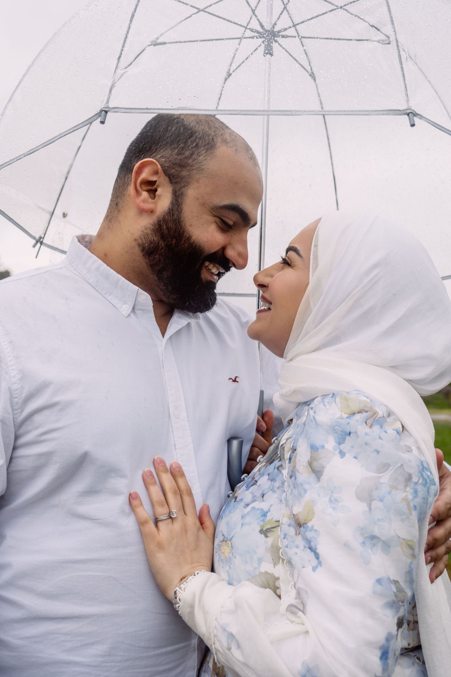 Happy couple sharing intimate moment under clear umbrella during pregnancy photoshoot.