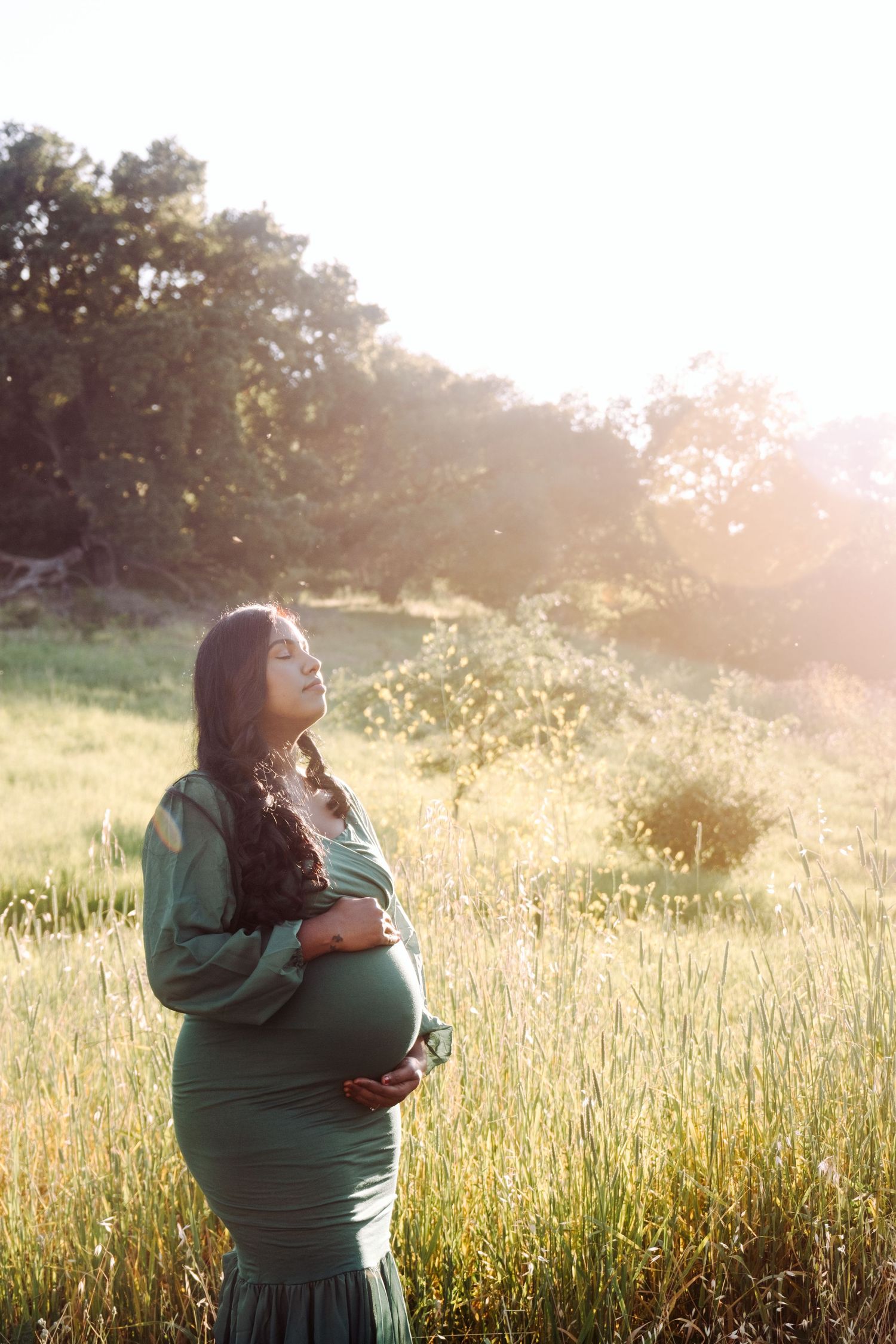 Silhouette of pregnant woman in green dress standing in sunlit field.