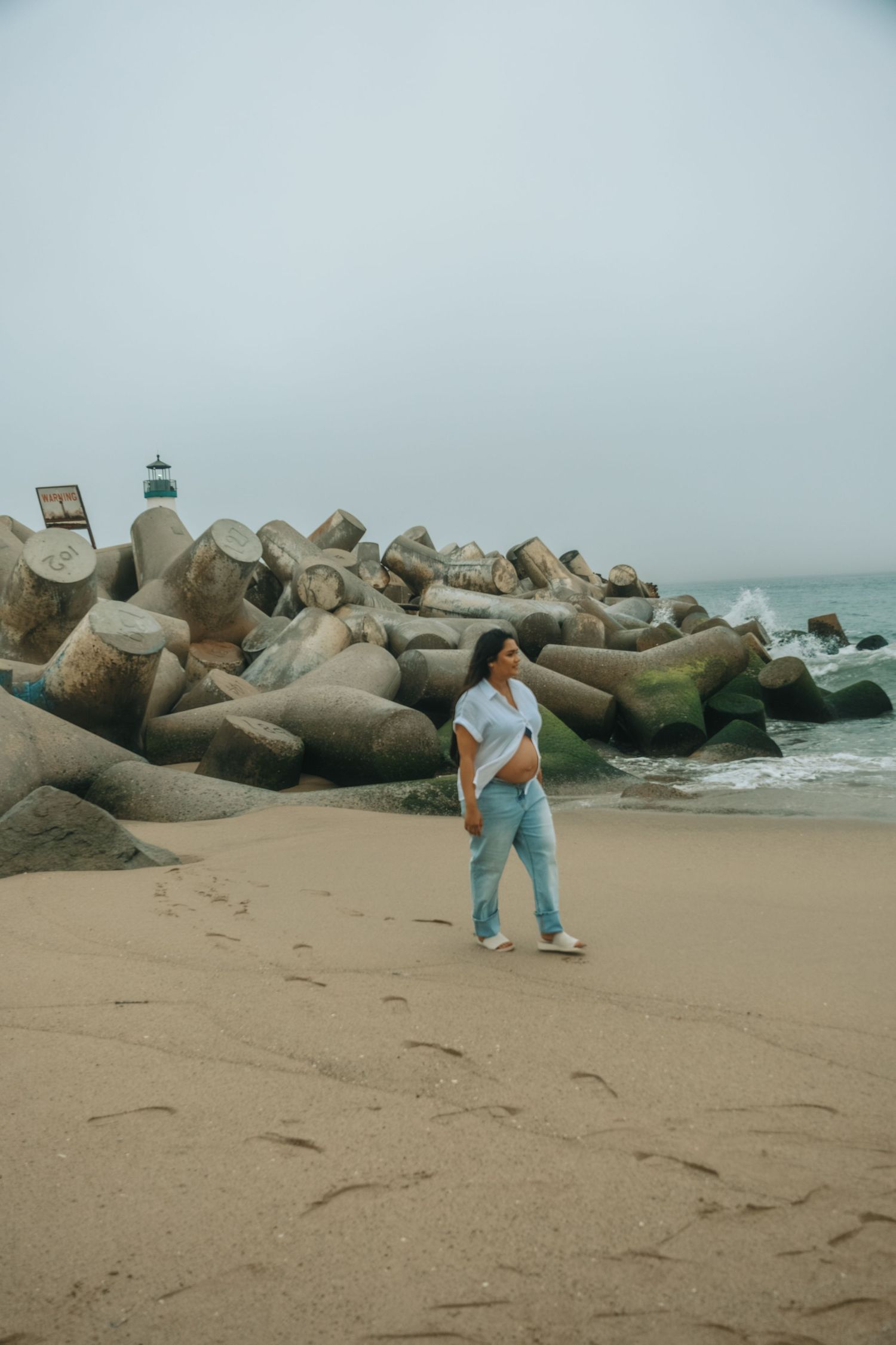 Figure in blue walks along a rocky beach with large concrete breakwater blocks.