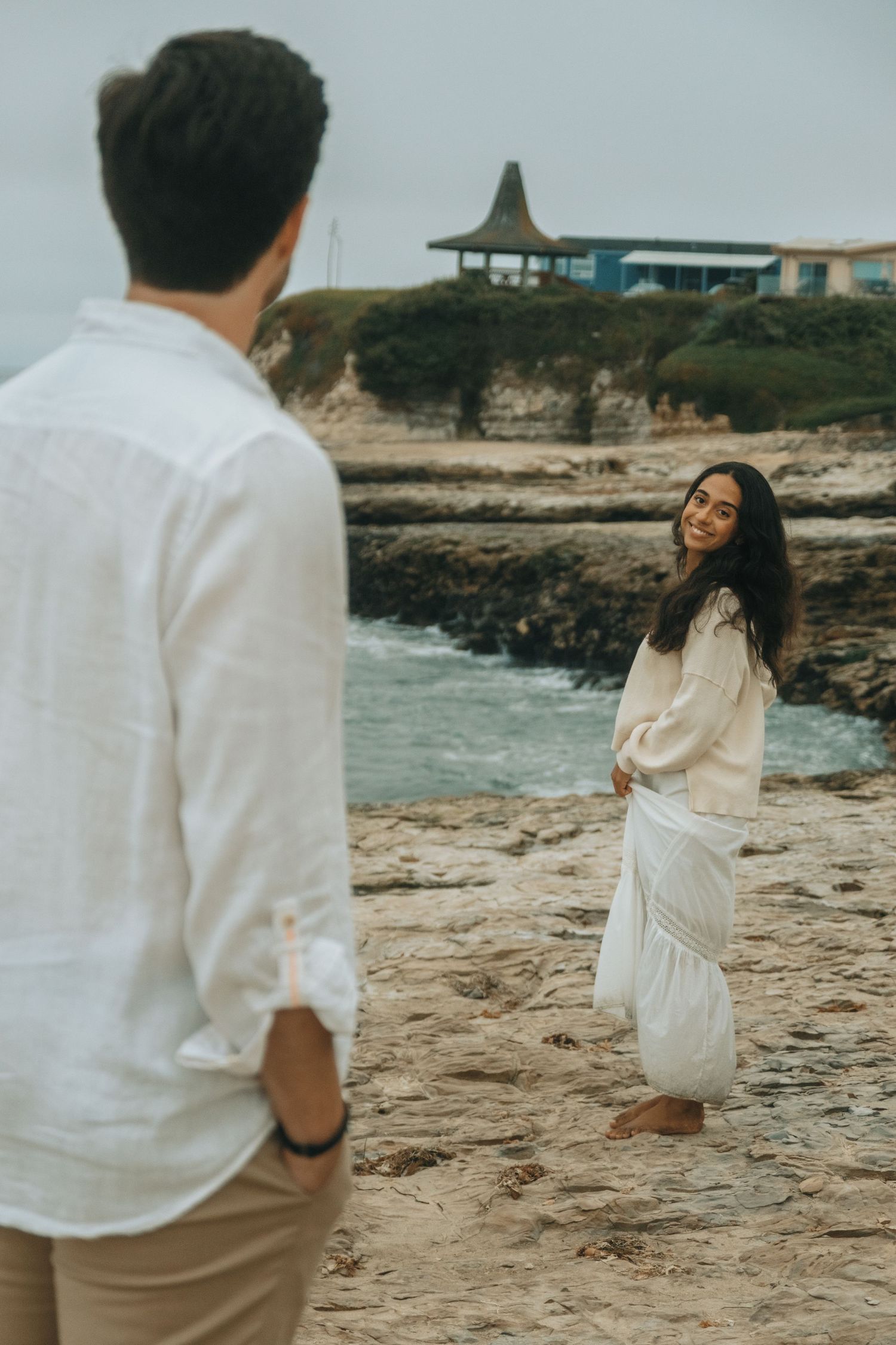 Couple in white clothing enjoying playful moment on rocky beach landscape.