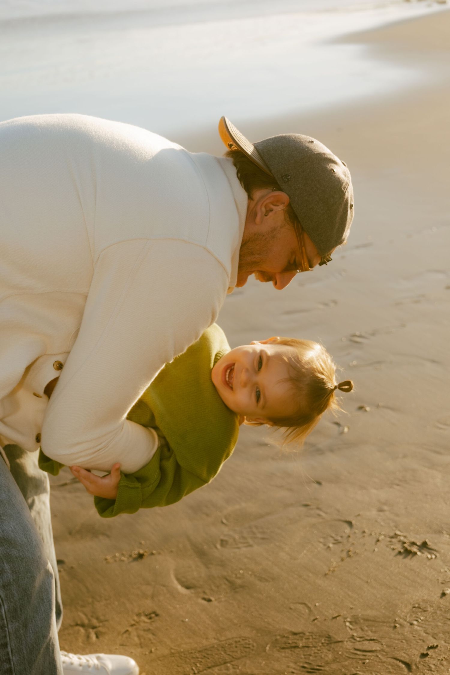 Adult in cap playfully swinging smiling toddler in green sweater on sandy beach at golden hour.