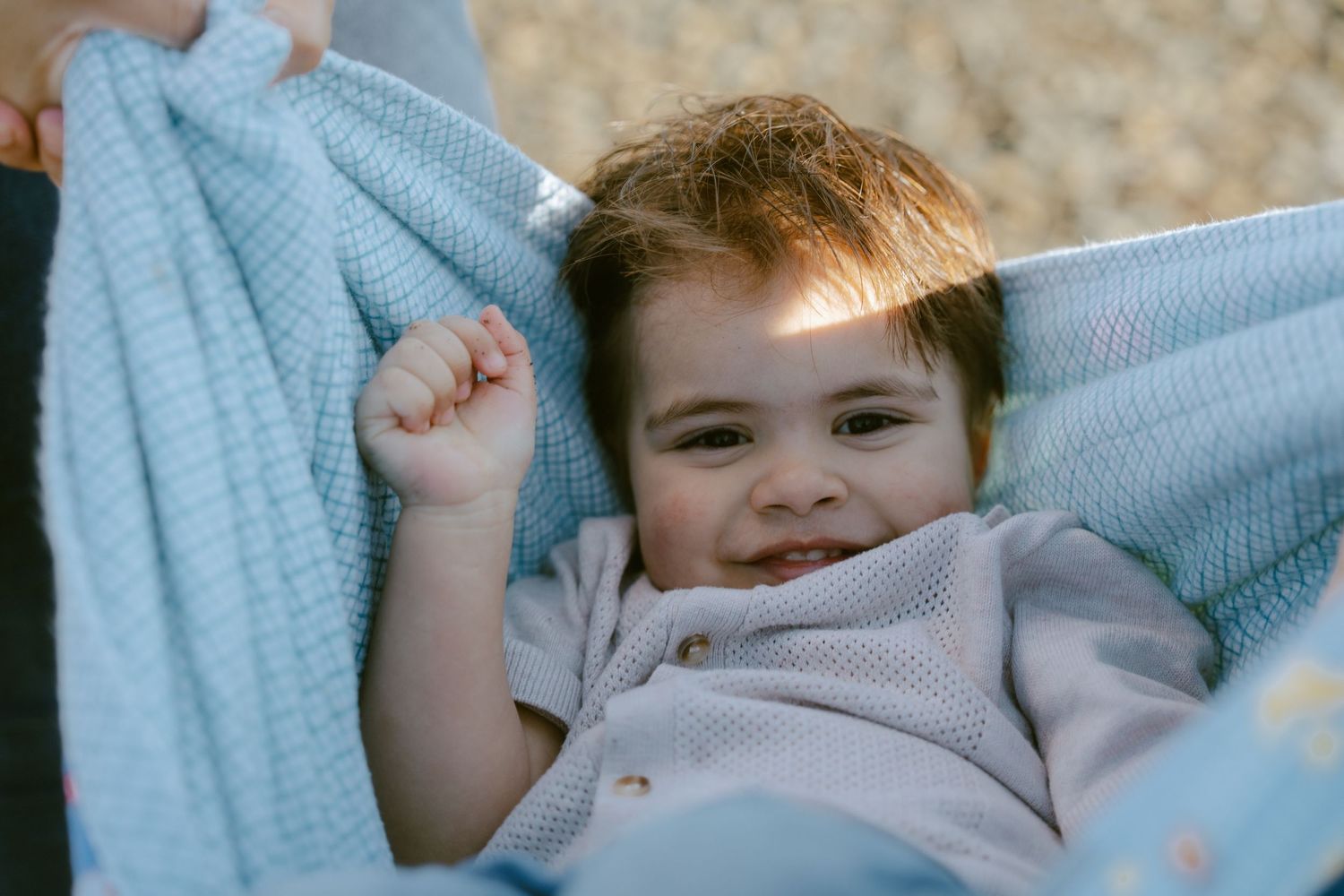 Smiling baby boy lying in a light blue knit blanket, raising his fist with a joyful expression outdoors.