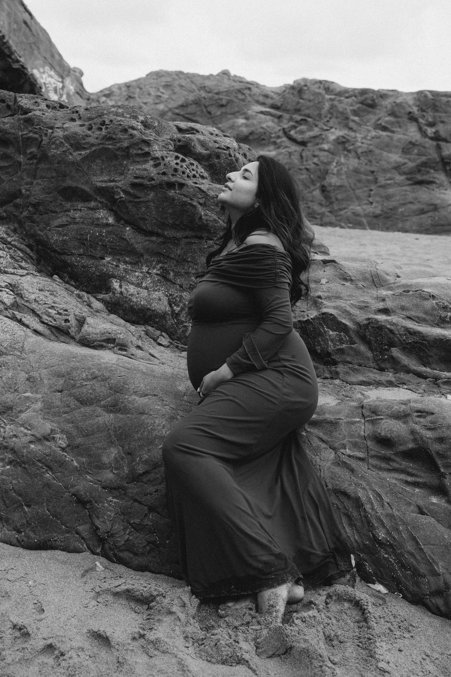 Black and white photo of a pregnant woman in a long dress posing against rocky cliffs on a beach.