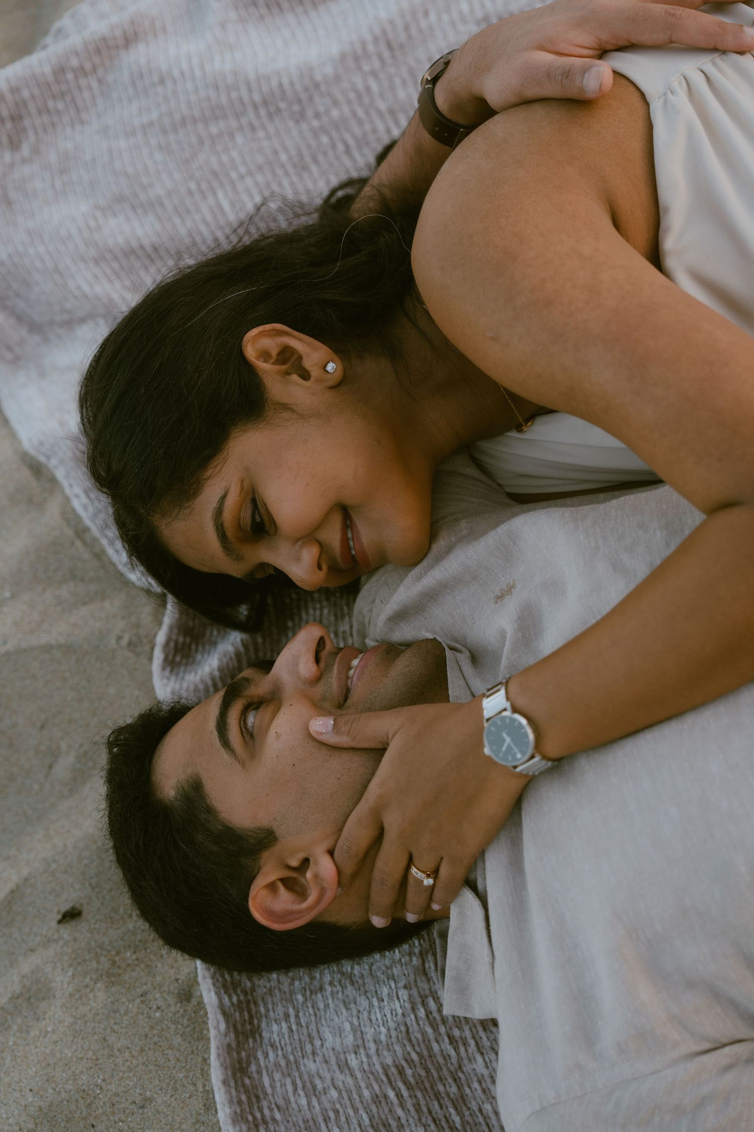 A couple shares a tender moment lying on a beach blanket, with the woman leaning over the man, both smiling warmly.