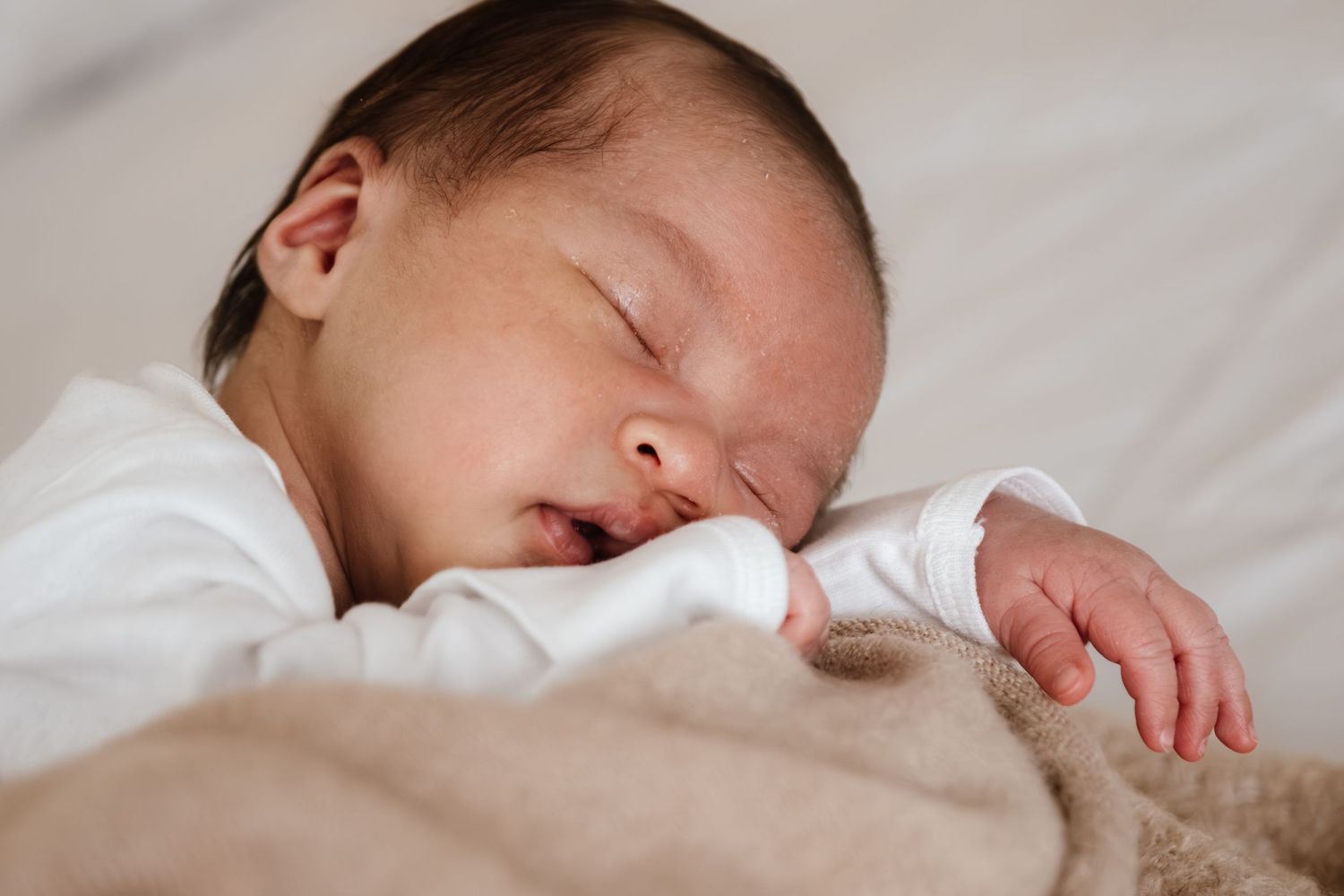 Peaceful newborn baby sleeping on a soft beige blanket, wearing a white onesie with mouth slightly open.