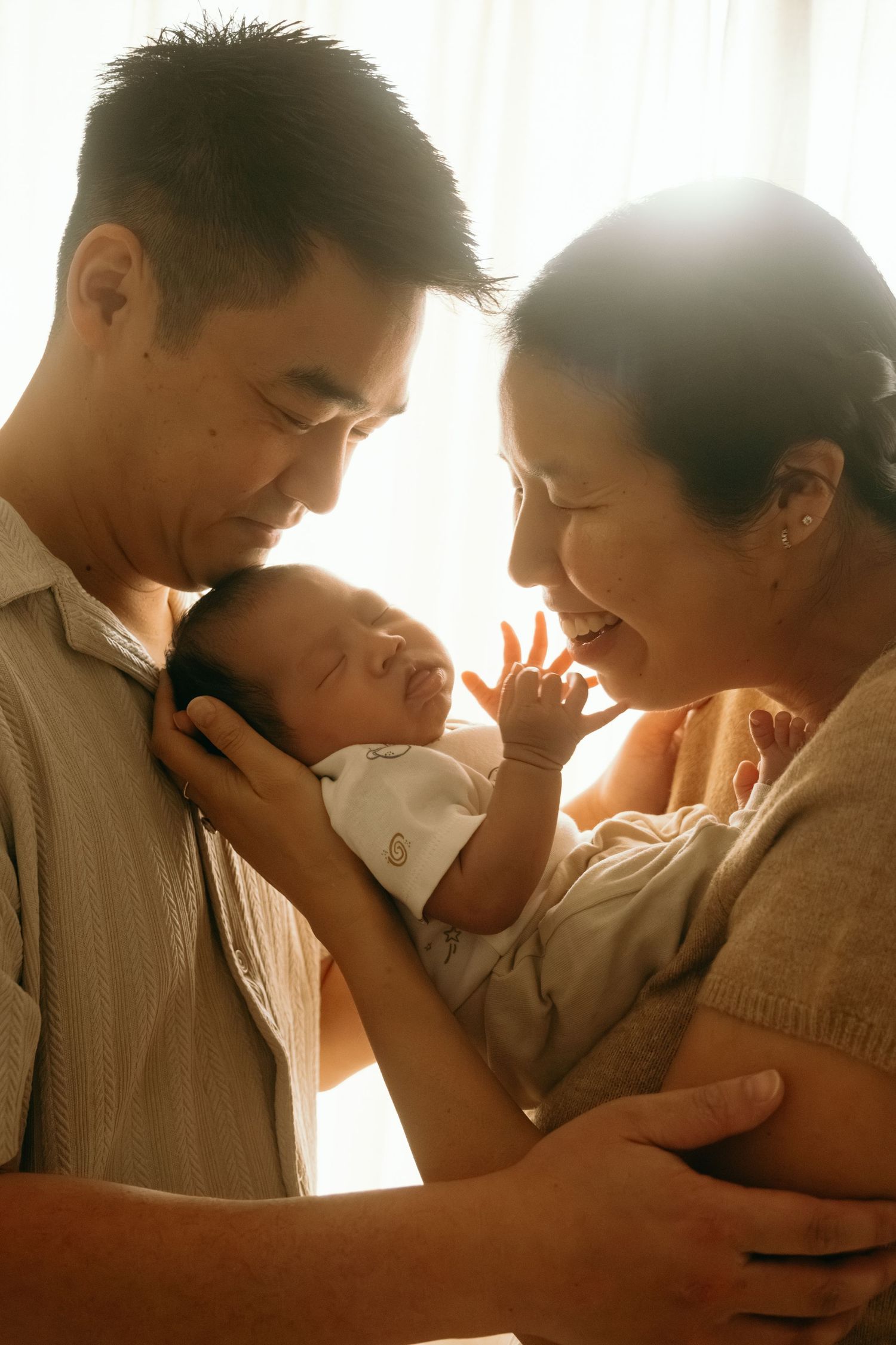 Asian parents smiling and bonding with their newborn baby in warm glowing natural light at home.