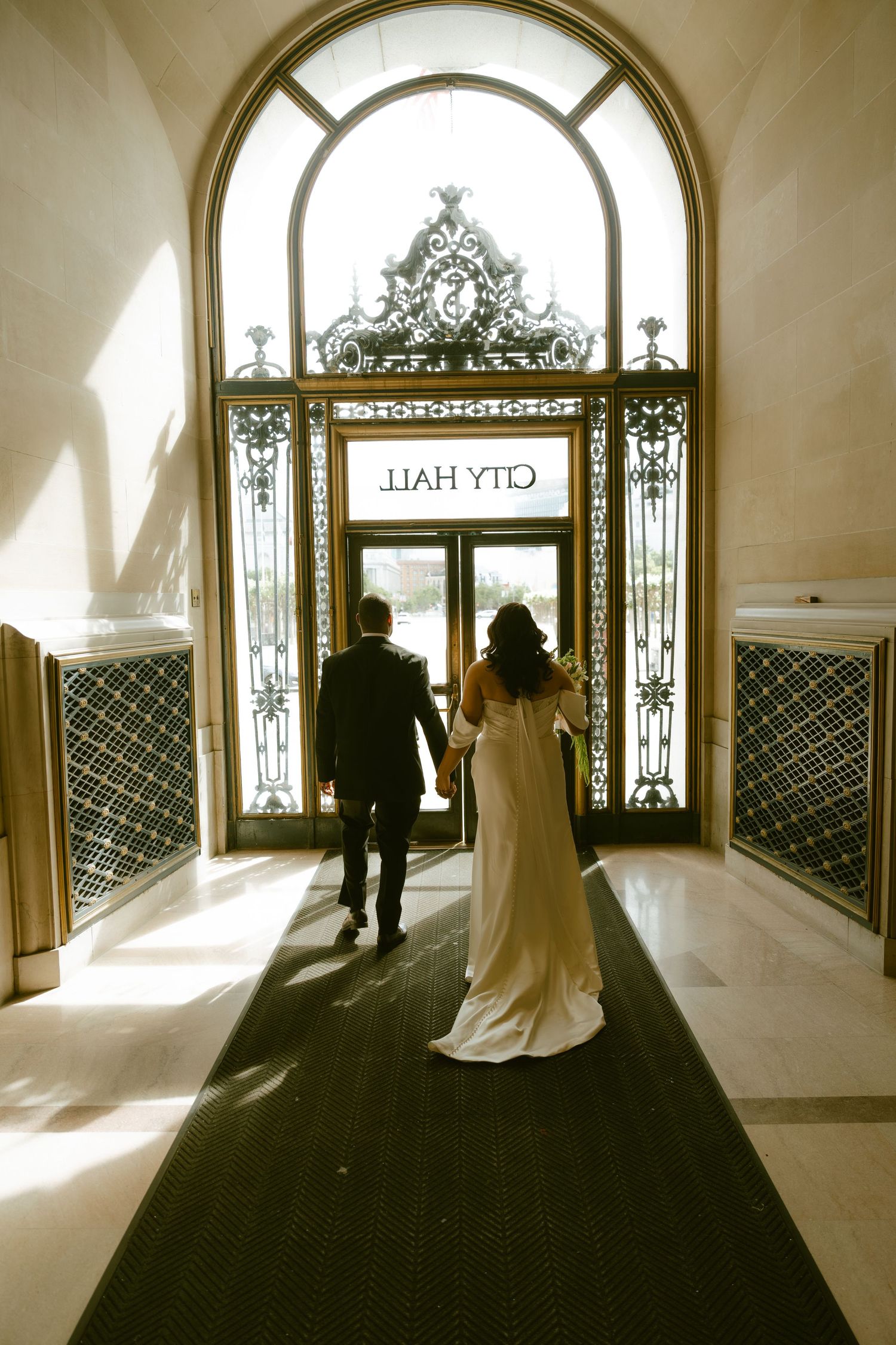 Bride and groom walking hand in hand toward ornate City Hall entrance doors on their wedding day.
