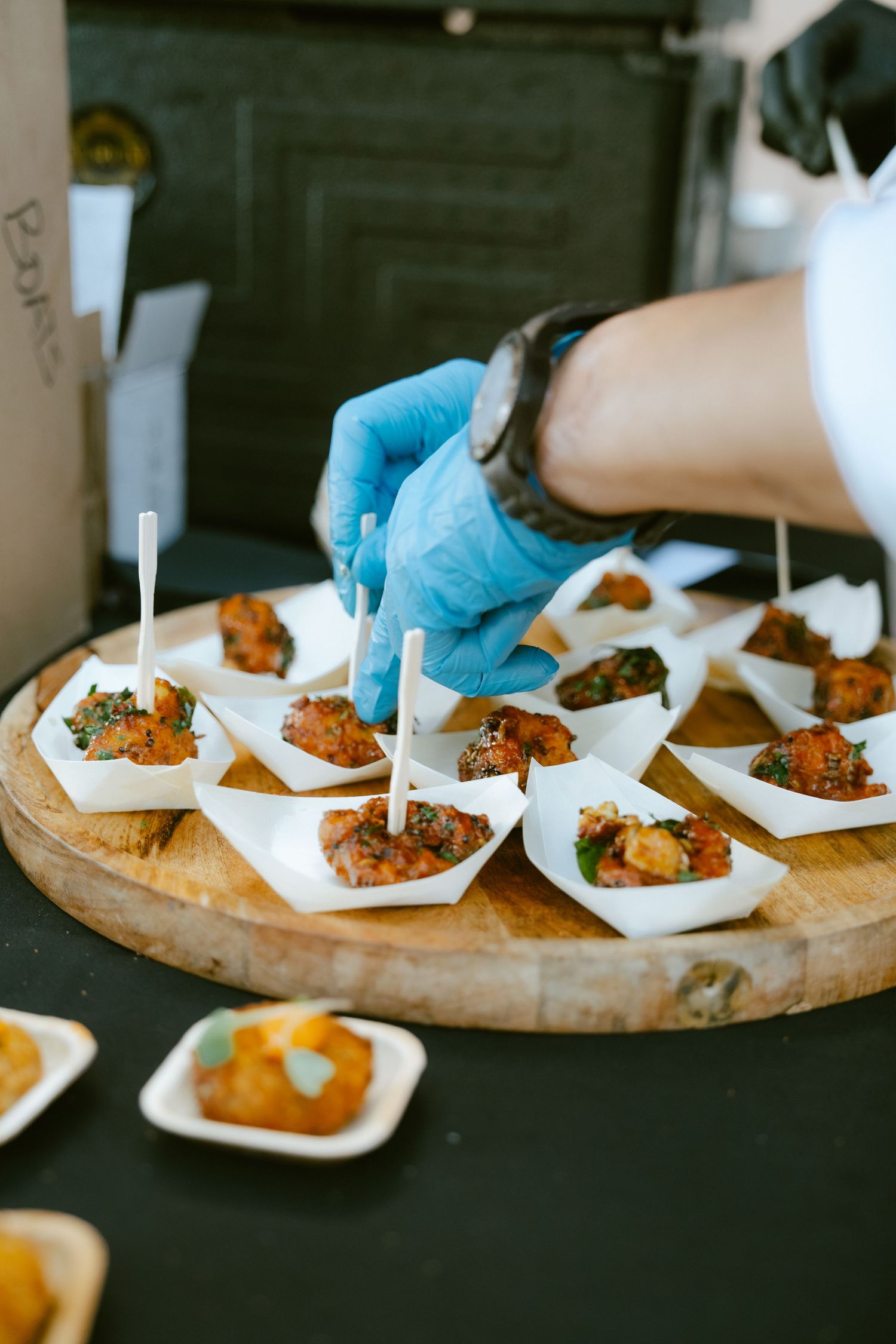 Chef in blue gloves arranging appetizers on white boats with toothpicks on a wooden serving board.