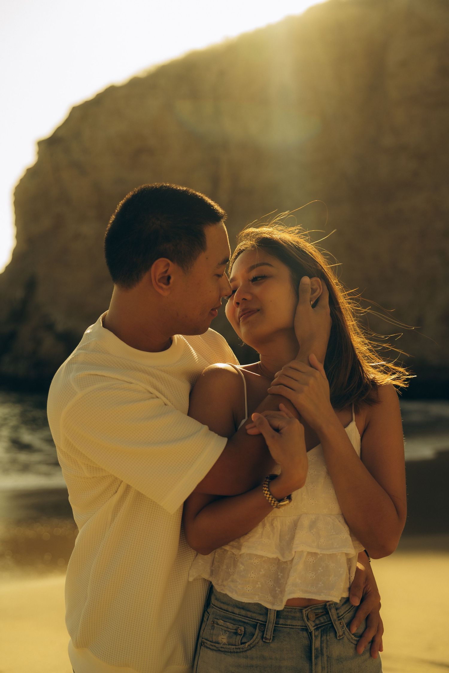 Couple sharing a romantic moment at sunset on a beach, with golden light and rocky cliffs in the background.
