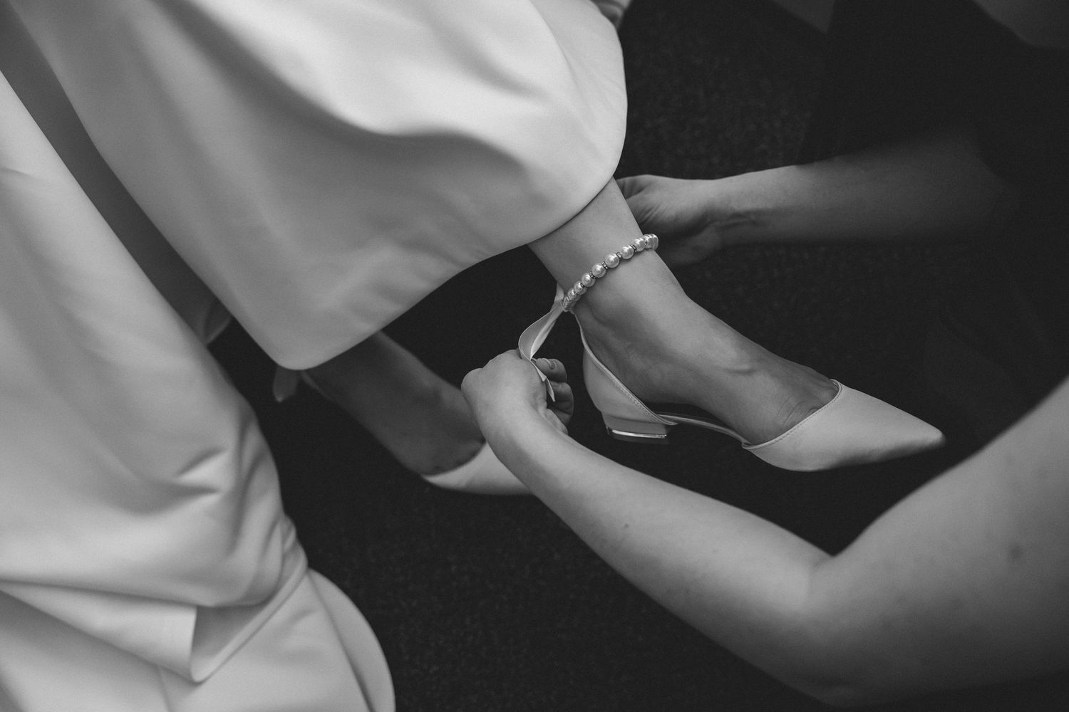 Black and white photo of a bridesmaid helping a bride put on a pearl anklet and heeled wedding shoe.