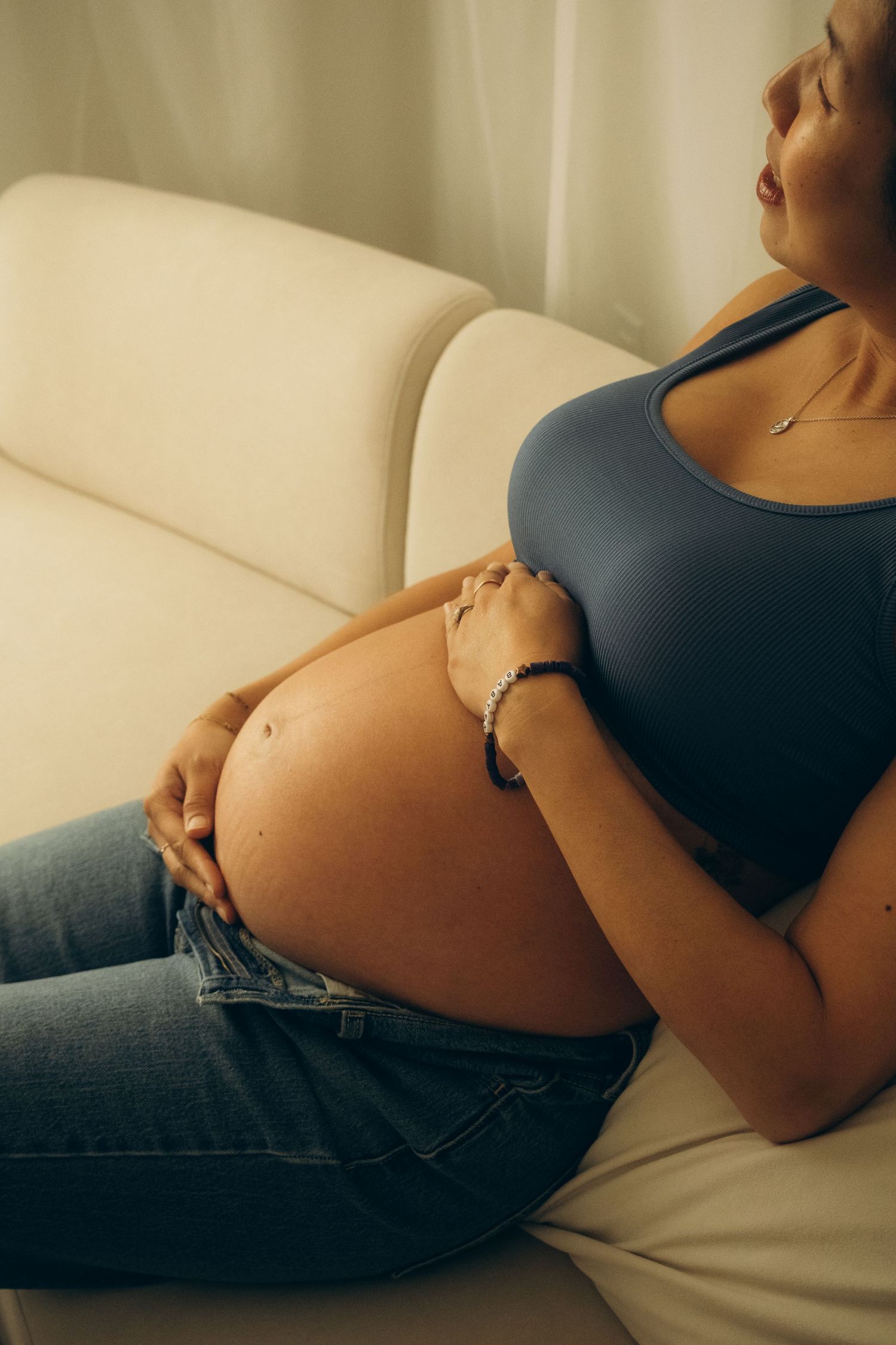 Expectant mother relaxing on white couch wearing blue top and jeans.