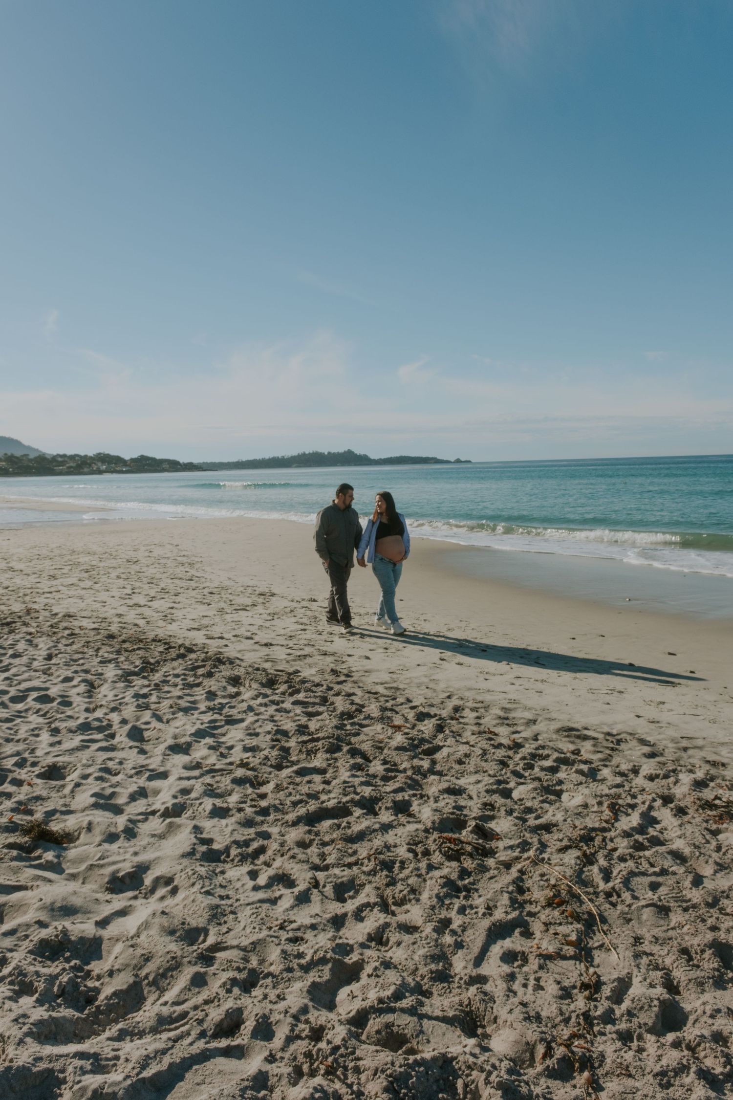 A couple walks along a scenic beach with waves and sand stretching into the distance.