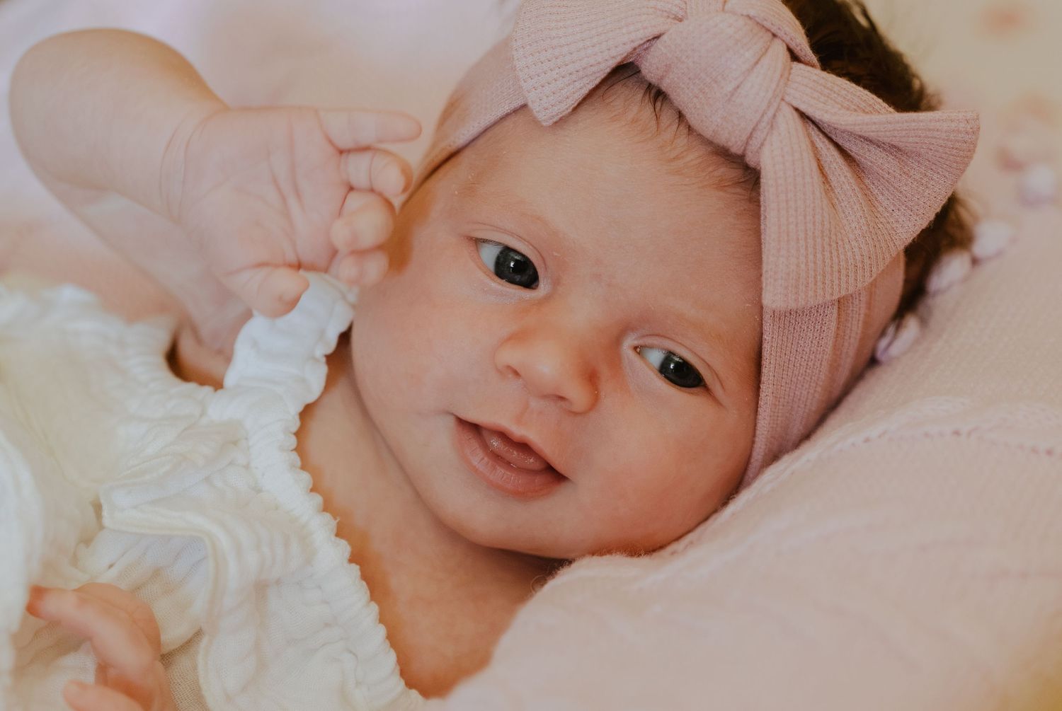 Newborn baby girl wearing a pink bow headband and white ruffled outfit, lying on a soft pillow.
