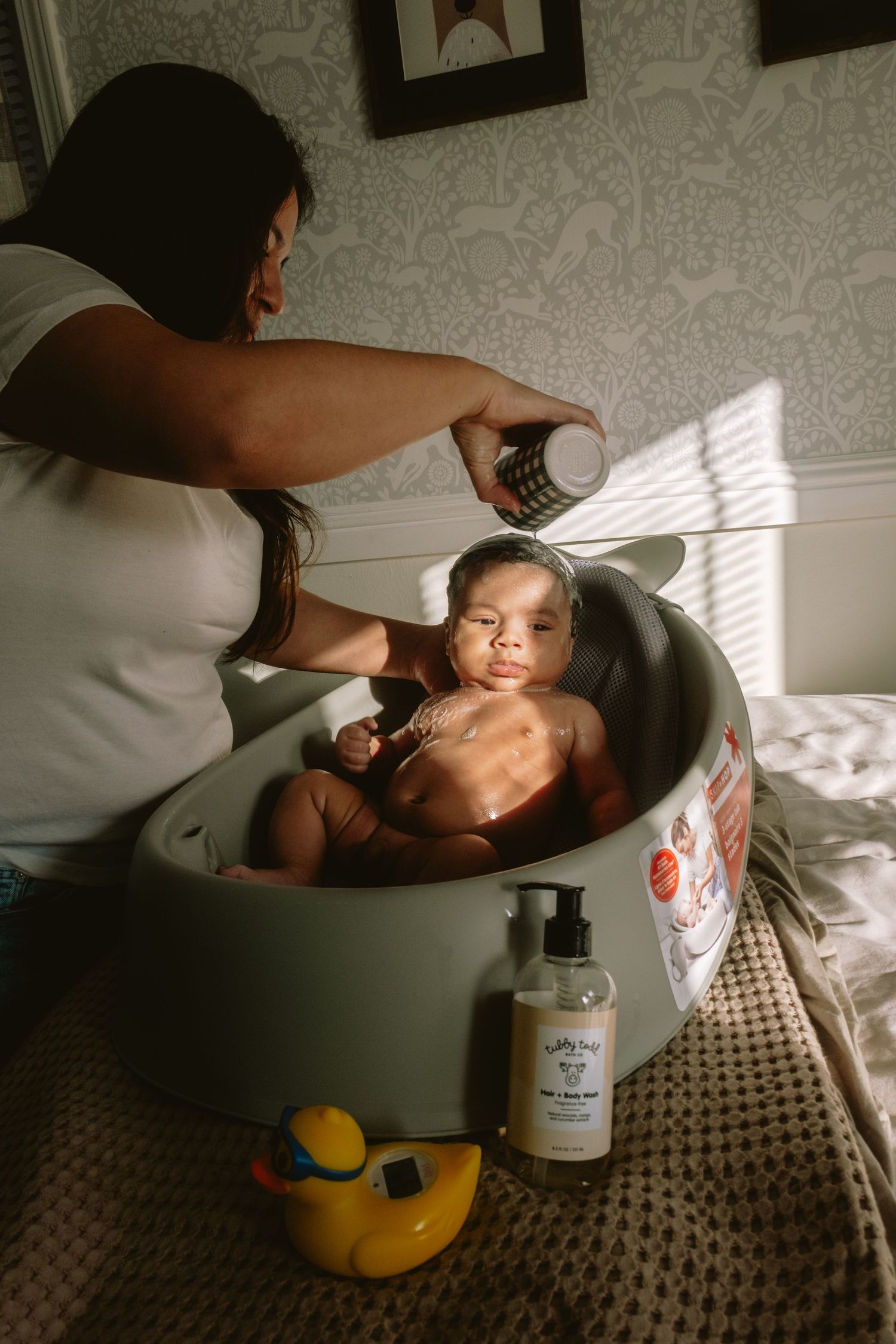 Mother bathing smiling baby in round tub with rubber duck and baby wash nearby in sunlit room.