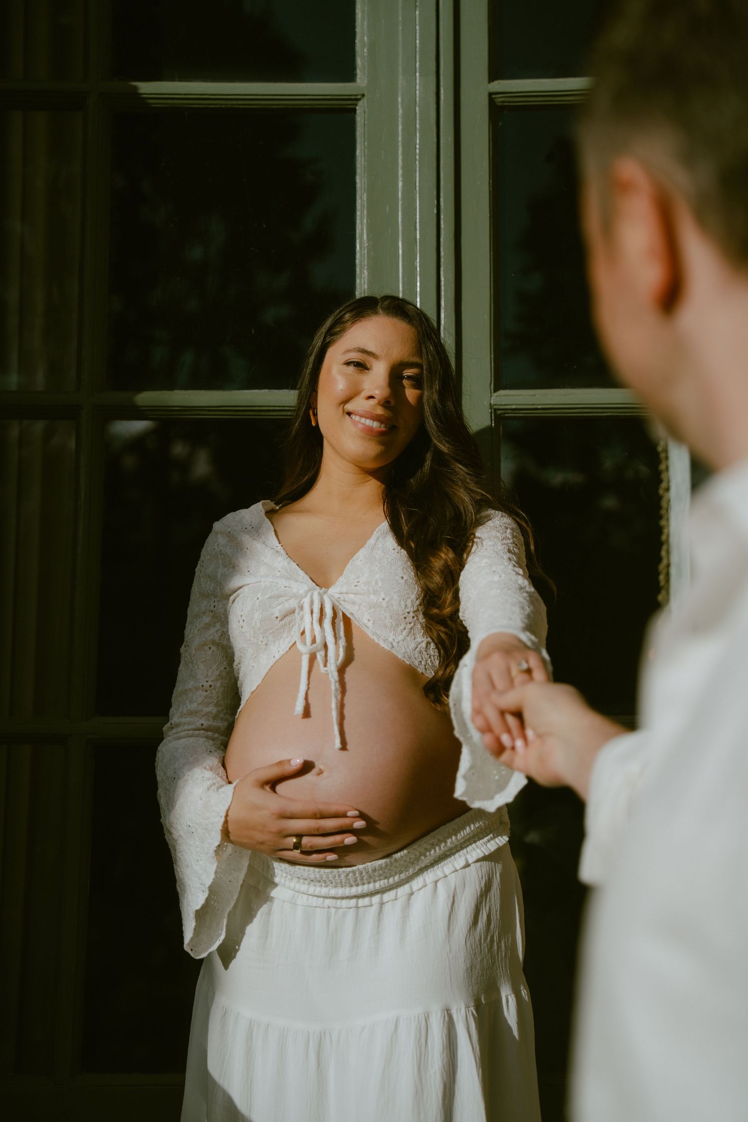 Smiling pregnant woman in white lace crop top holds baby bump while holding hands with partner near large window.