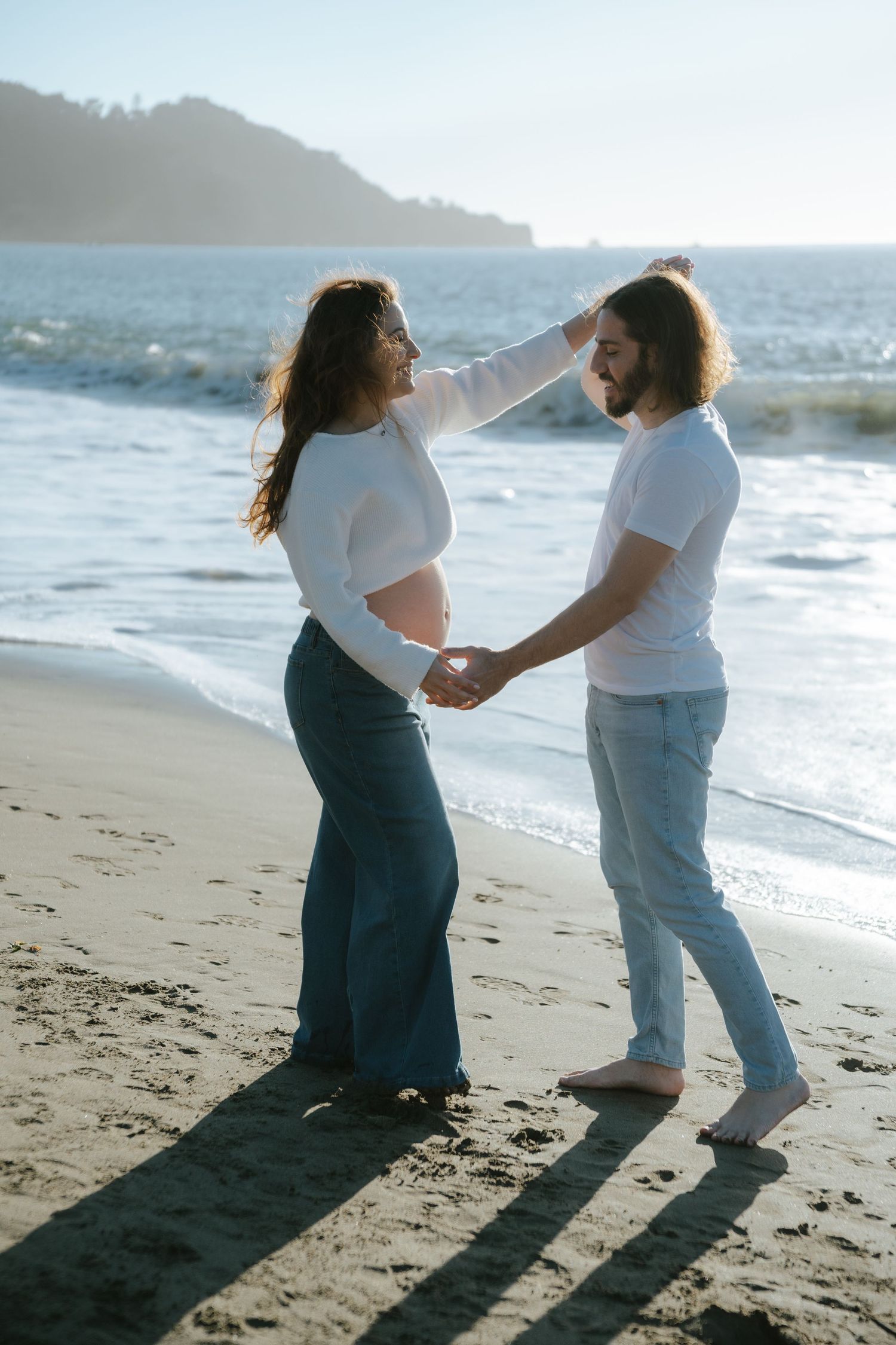 Pregnant couple holding hands on a sunny beach, woman touching man's hair, wearing white tops and jeans.