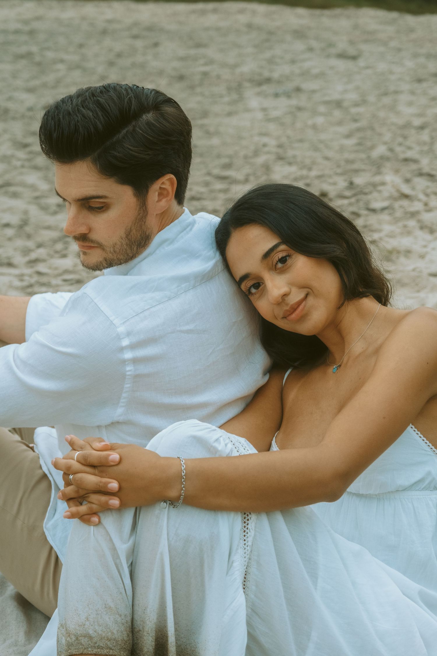 Couple in white sitting on sandy beach, woman smiling and leaning on mans back holding hands.