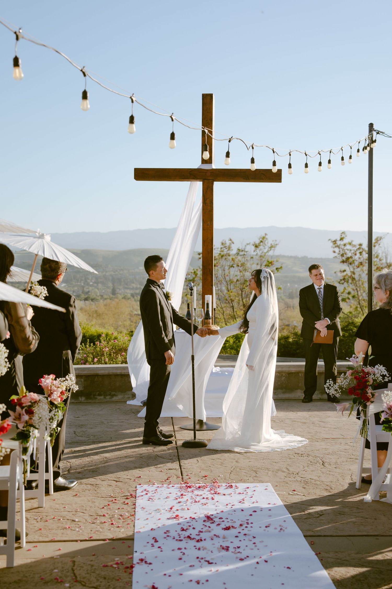 Outdoor wedding ceremony with couple at wooden cross altar, rose petal aisle, string lights, and mountain views.