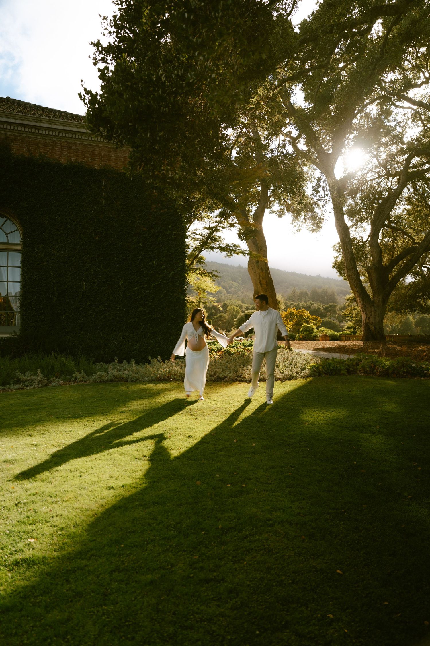 Couple in white outfits holding hands on lush green lawn beside ivy-covered building with sunlight filtering through trees.