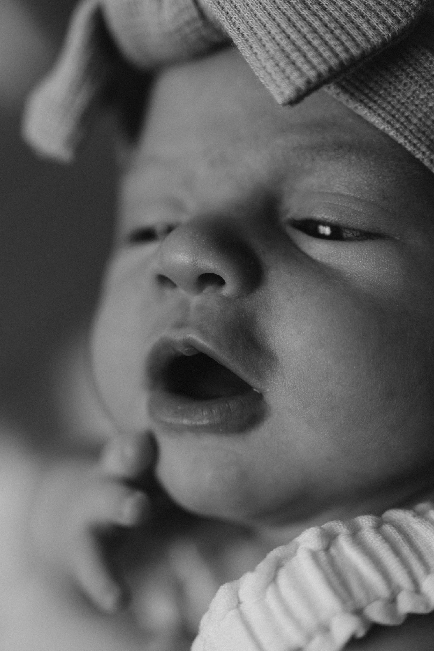 Black and white close-up of a newborn baby slightly opening eyes, wearing a knit hat and striped outfit.