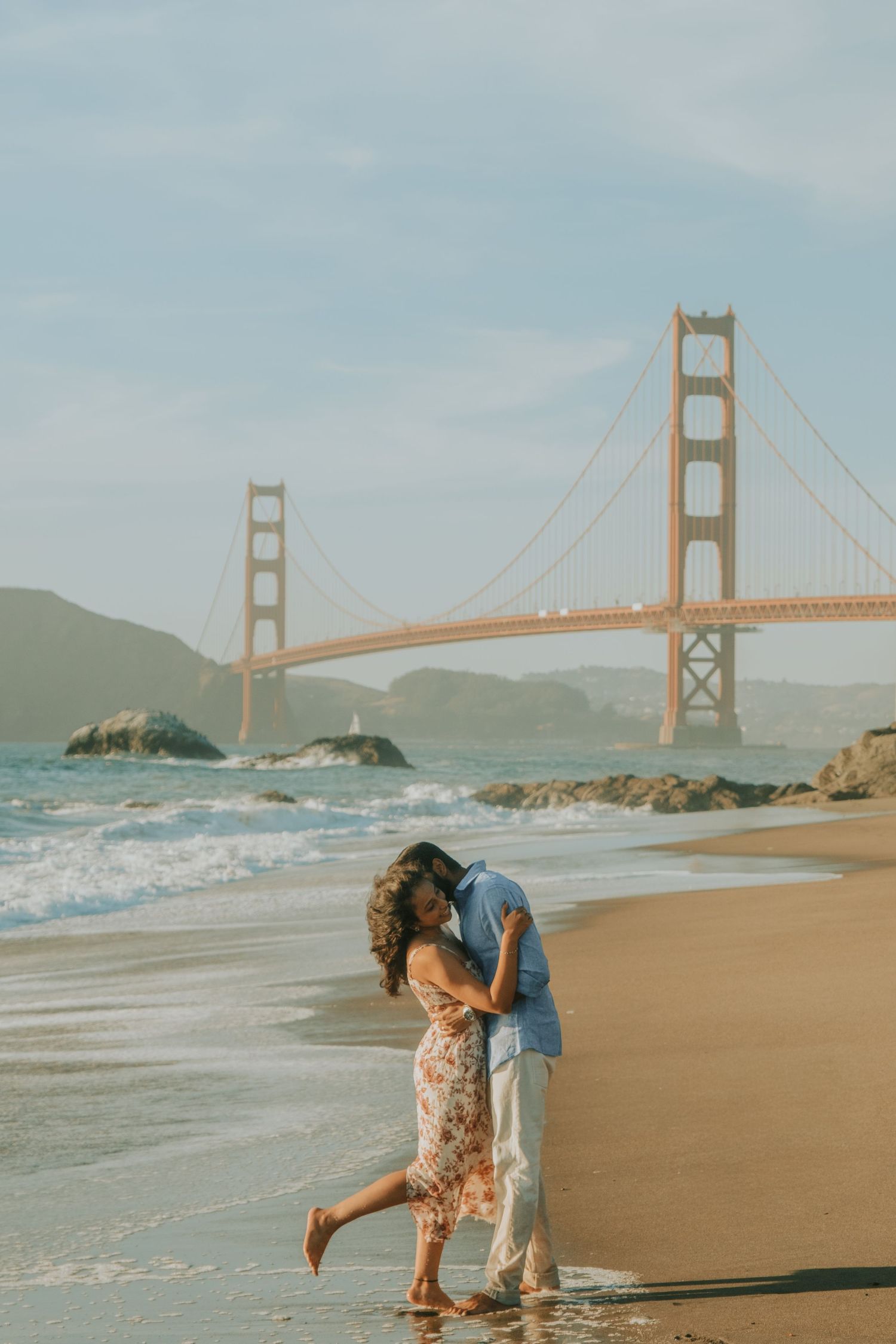 A couple dancing playfully on Baker Beach with the Golden Gate Bridge in the distance.