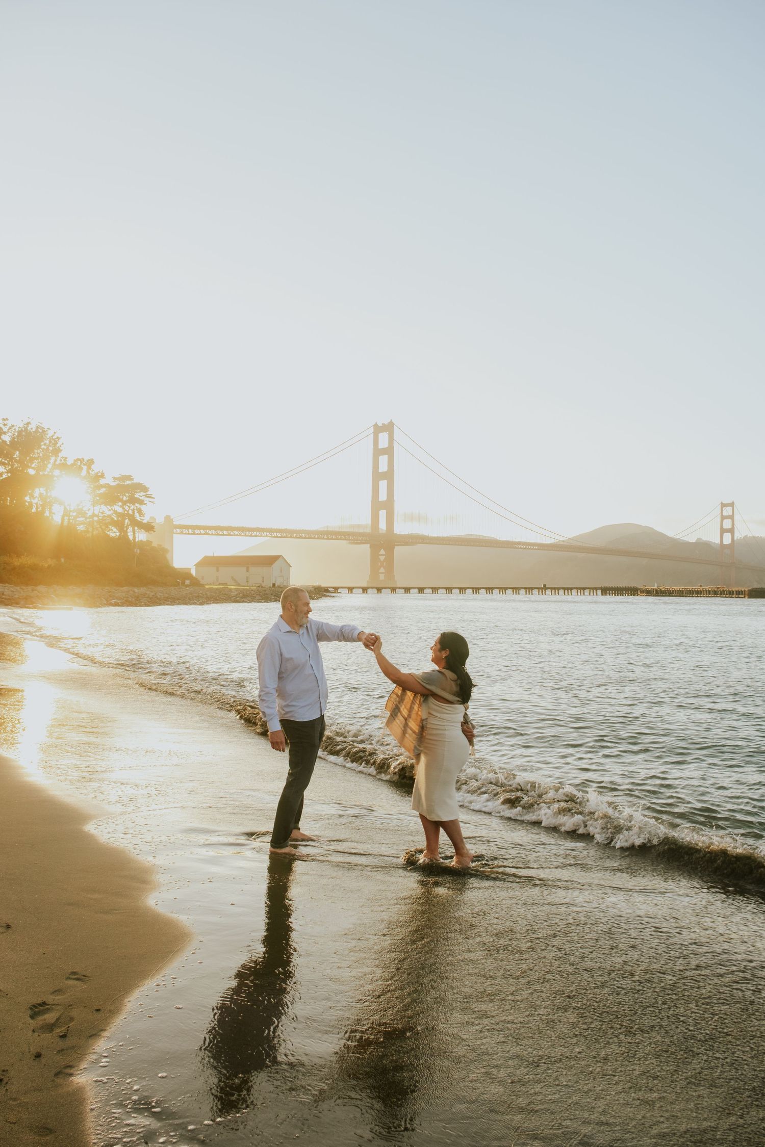 Couple plays in waves at beach with Golden Gate Bridge silhouetted in background at sunset.