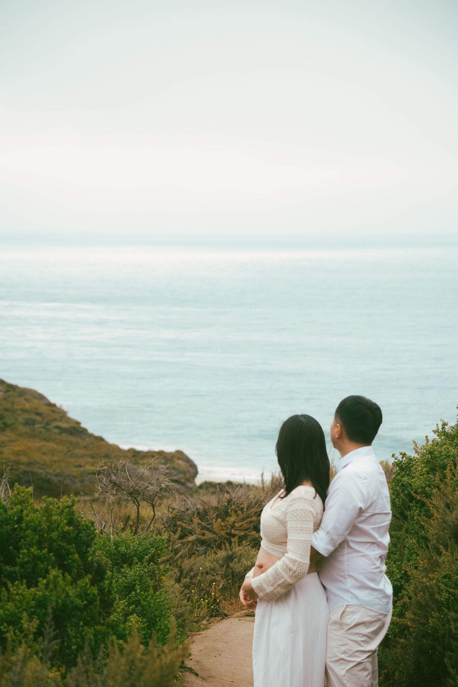 A couple in white clothing stands on a coastal path overlooking the ocean.