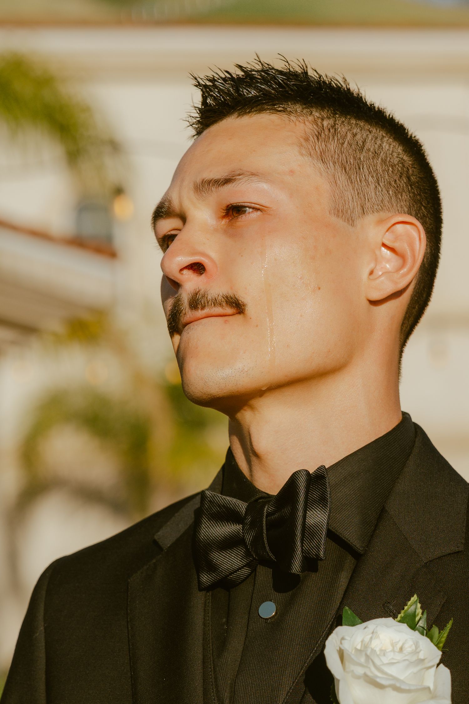 Groom in black tuxedo with bow tie and white rose boutonniere, looking away thoughtfully outdoors.