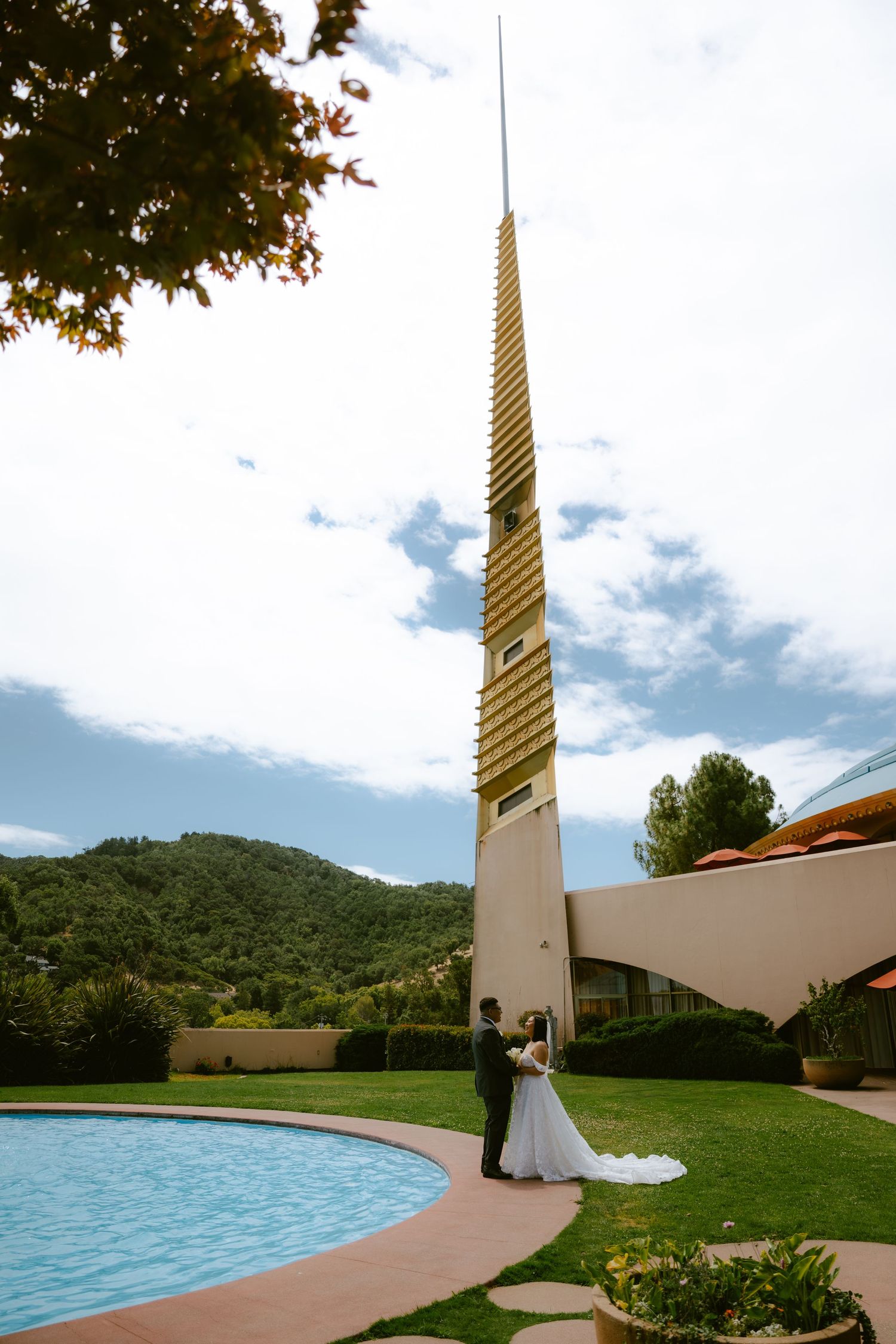 Wedding couple standing by pool beneath tall golden spire tower with mountain backdrop on sunny day.
