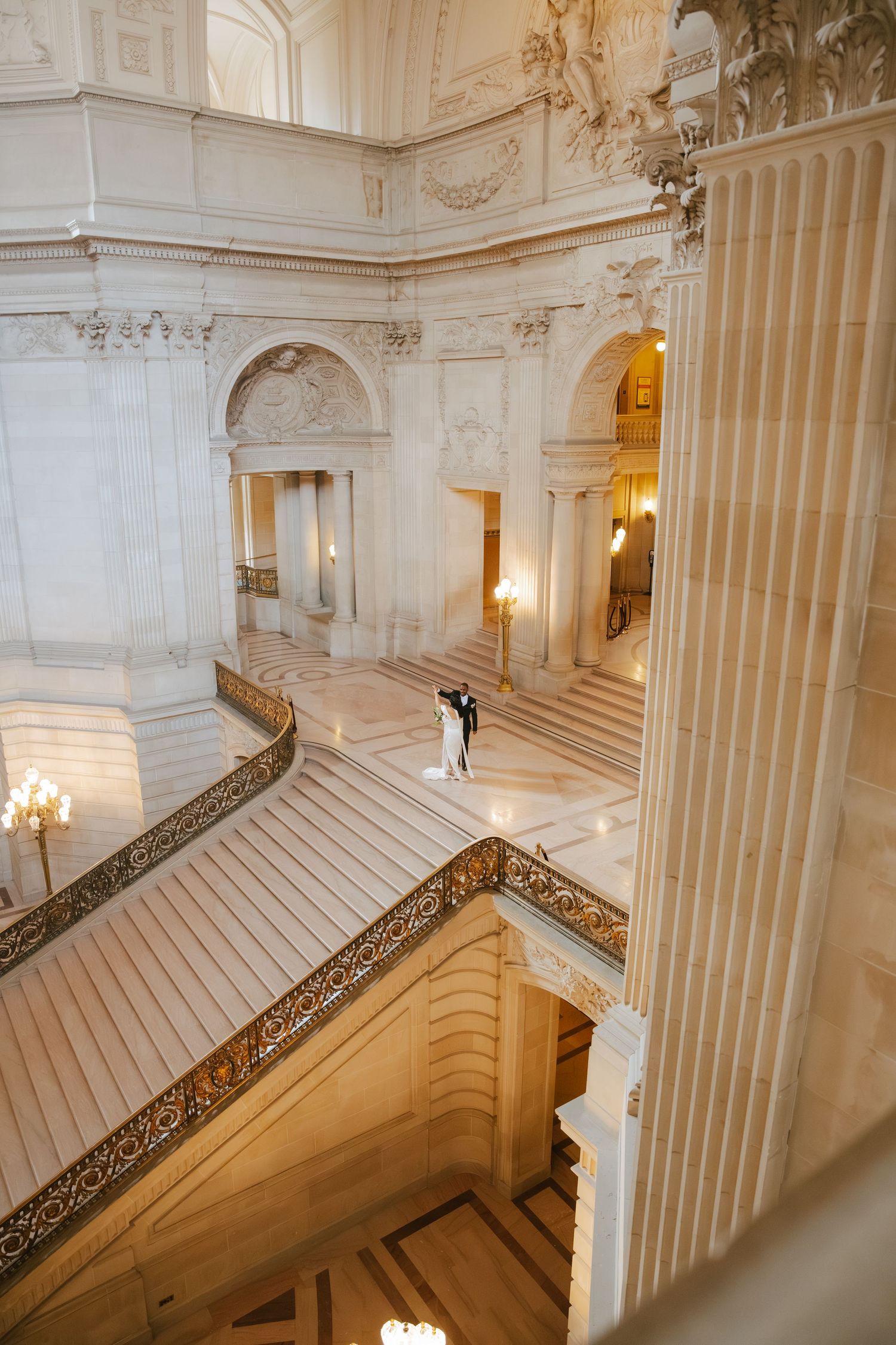 Bride and groom embrace on grand marble staircase inside San Francisco City Hall's ornate Beaux-Arts rotunda.